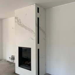Grzegorz Zegzula - Nowoczesny kominek obudowany białym marmurem z czarnymi akcentami, widoczne niedokończone prace elektryczne i surowy beton na podłodze.