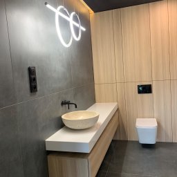 Grzegorz Zegzula - Nowoczesna łazienka z szarymi płytkami, drewnianą zabudową, kamienną umywalką i podwieszaną toaletą, oświetlona podwieszanymi lampami w kształcie okręgów.