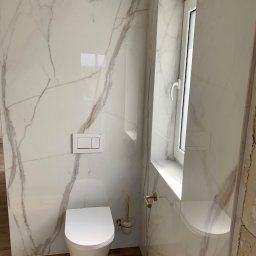 Grzegorz Zegzula - Zawieszona miska WC w nowoczesnej łazience wykończonej płytami imitującymi marmur Calacatta, drewnopodobna podłoga, złote akcenty w postaci uchwytu na szczotkę toaletową i relingu.