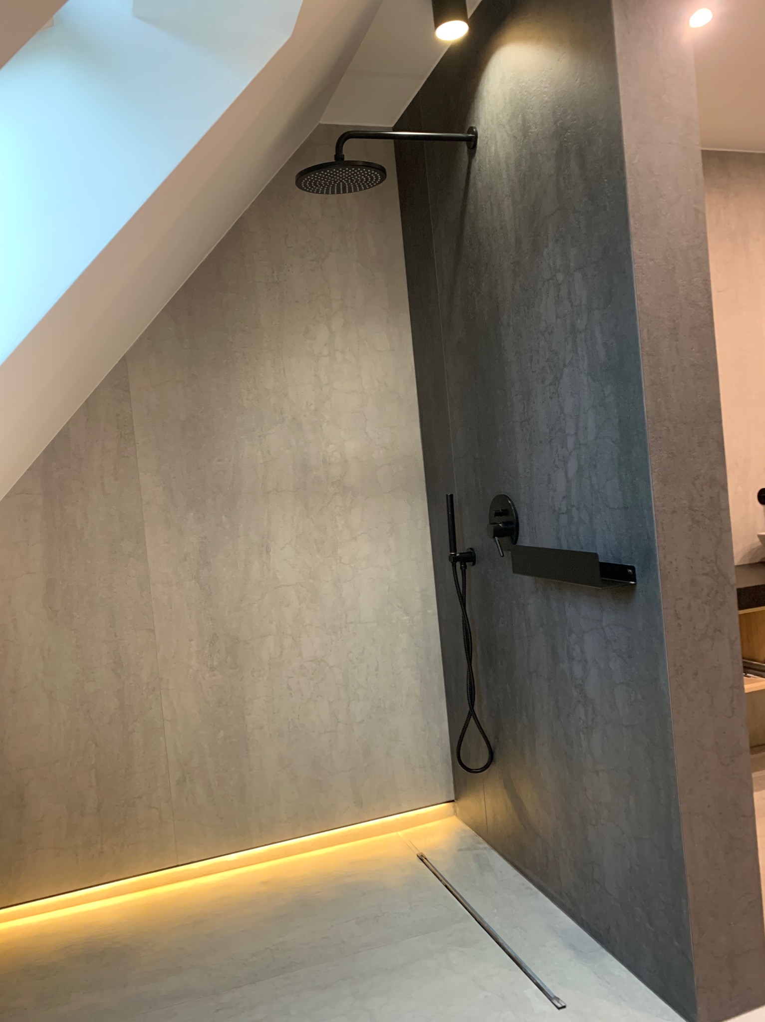 Nowoczesna kabina prysznicowa w łazience na poddaszu, wykończona płytkami imitującymi kamień w odcieniach szarości, z czarną deszczownicą, baterią i odpływem liniowym, podświetlona listwą LED przy...