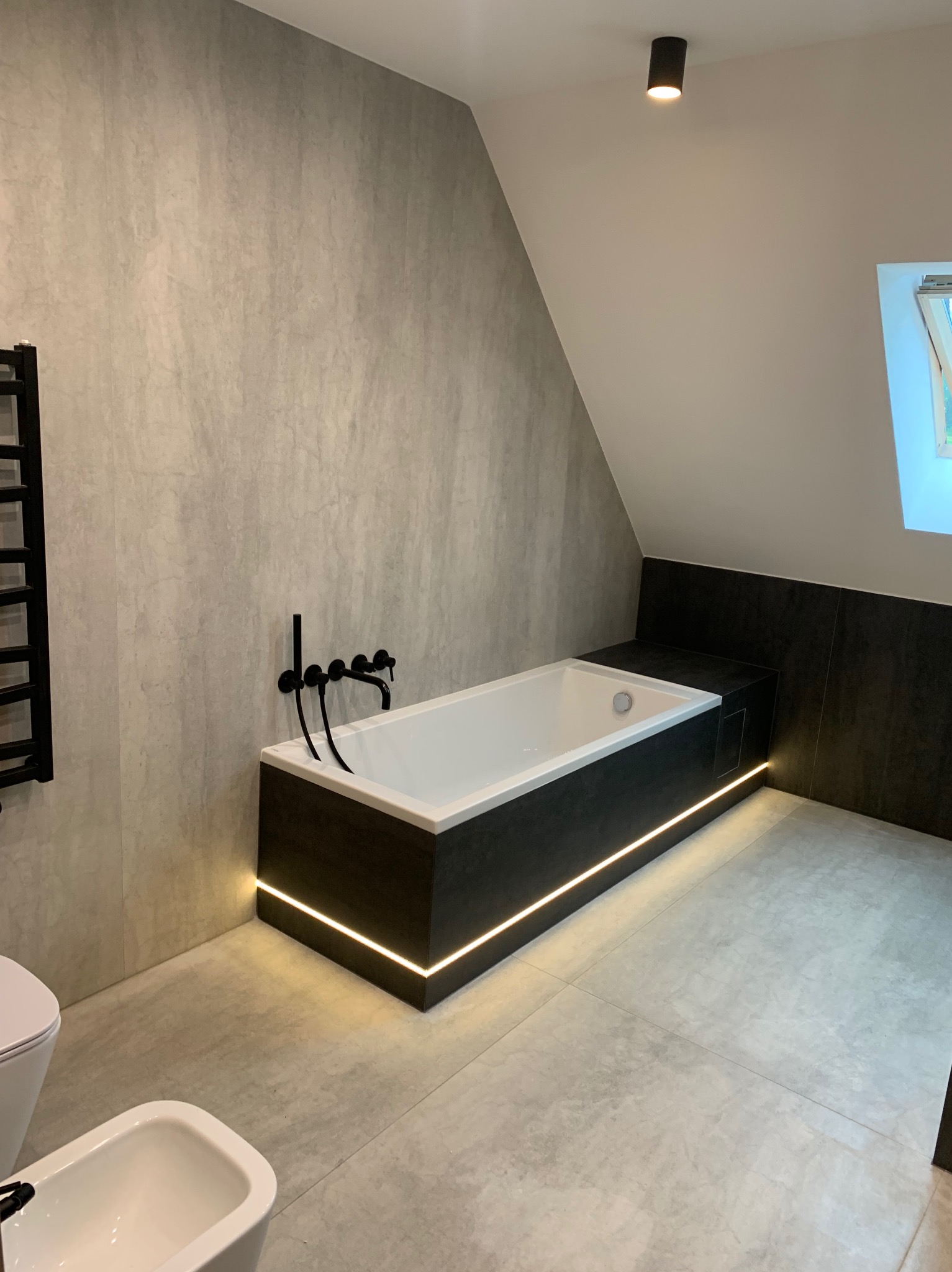 Nowoczesna łazienka na poddaszu: wanna obudowana ciemnoszarą płytą z podświetleniem LED, czarna armatura, szare płytki imitujące beton na ścianach i podłodze, grzejnik drabinkowy w kolorze czarnym,...