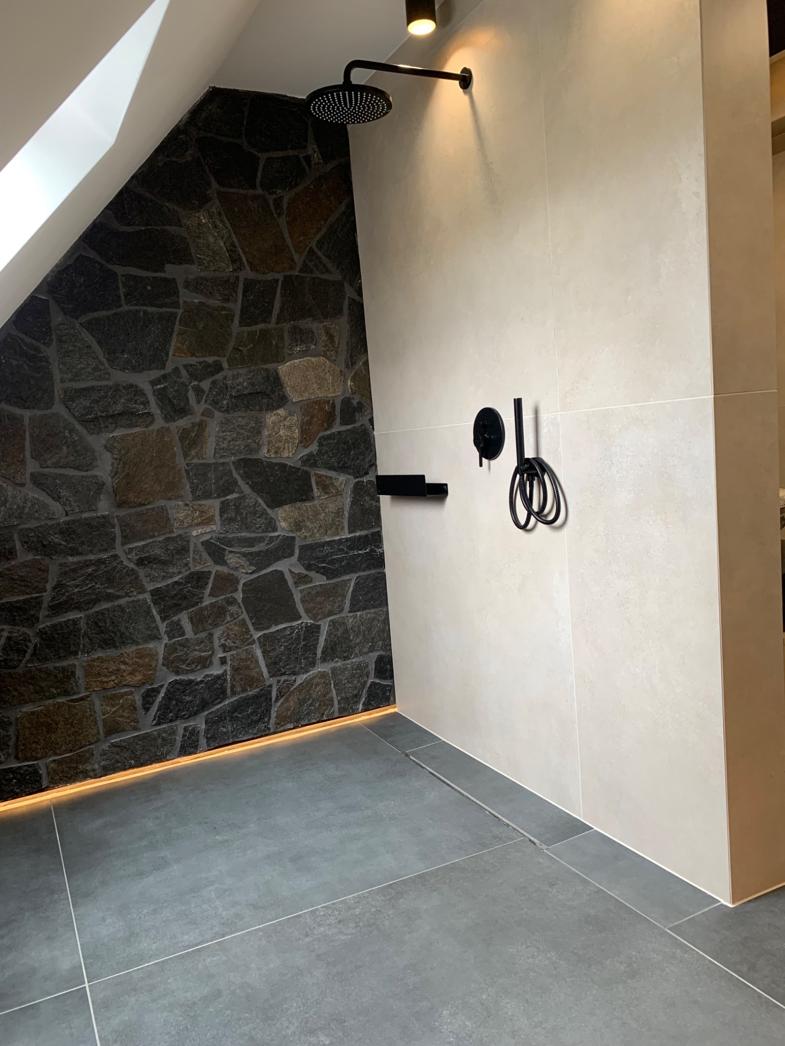 Nowoczesna kabina prysznicowa w łazience na poddaszu, ściana wyłożona nieregularnym kamieniem z podświetleniem LED, deszczownica i czarna armatura.
