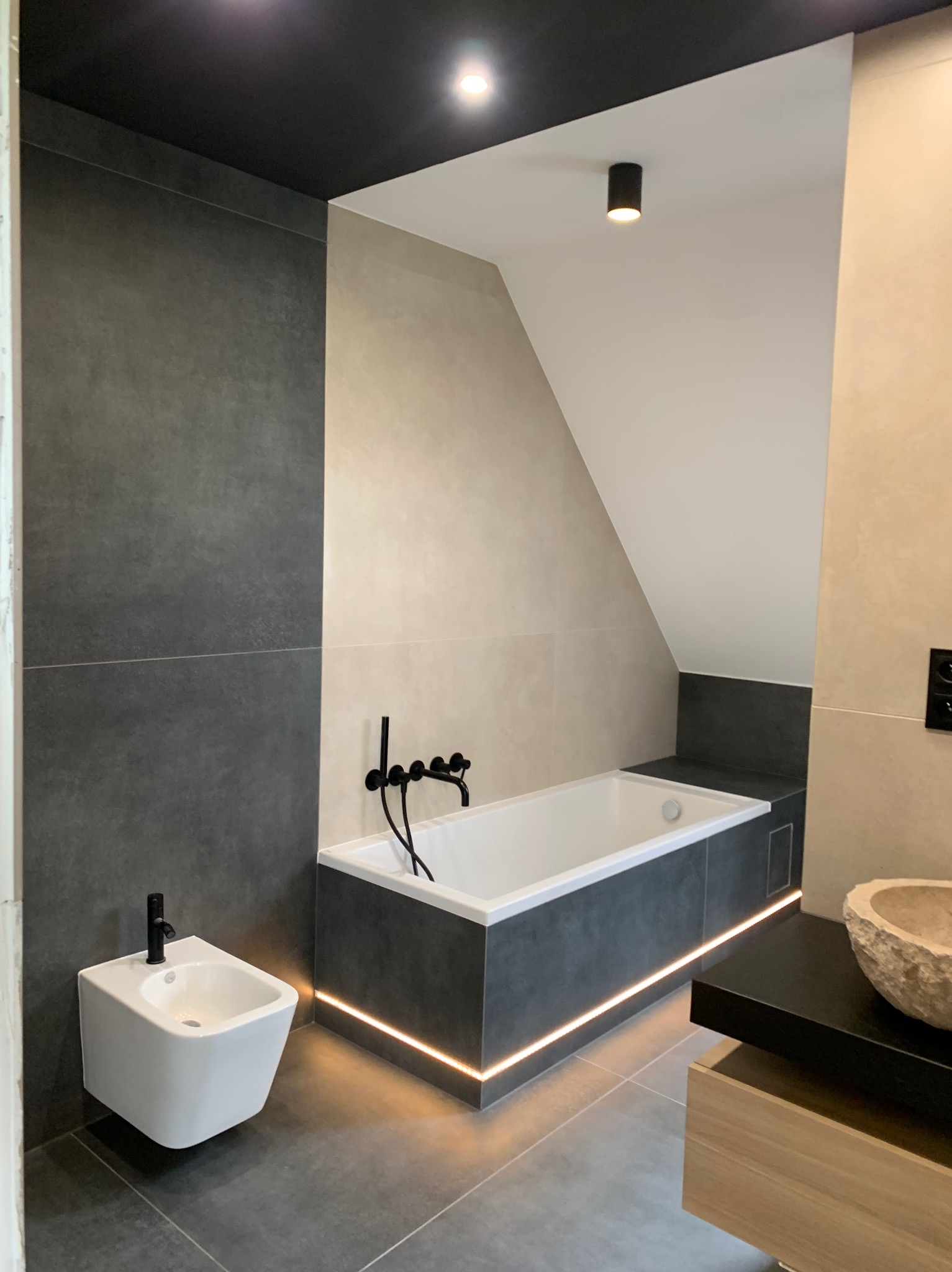 Nowoczesna łazienka w odcieniach szarości i beżu z podświetlaną wanną, wiszącym bidetem i umywalką w kształcie kamiennej misy.