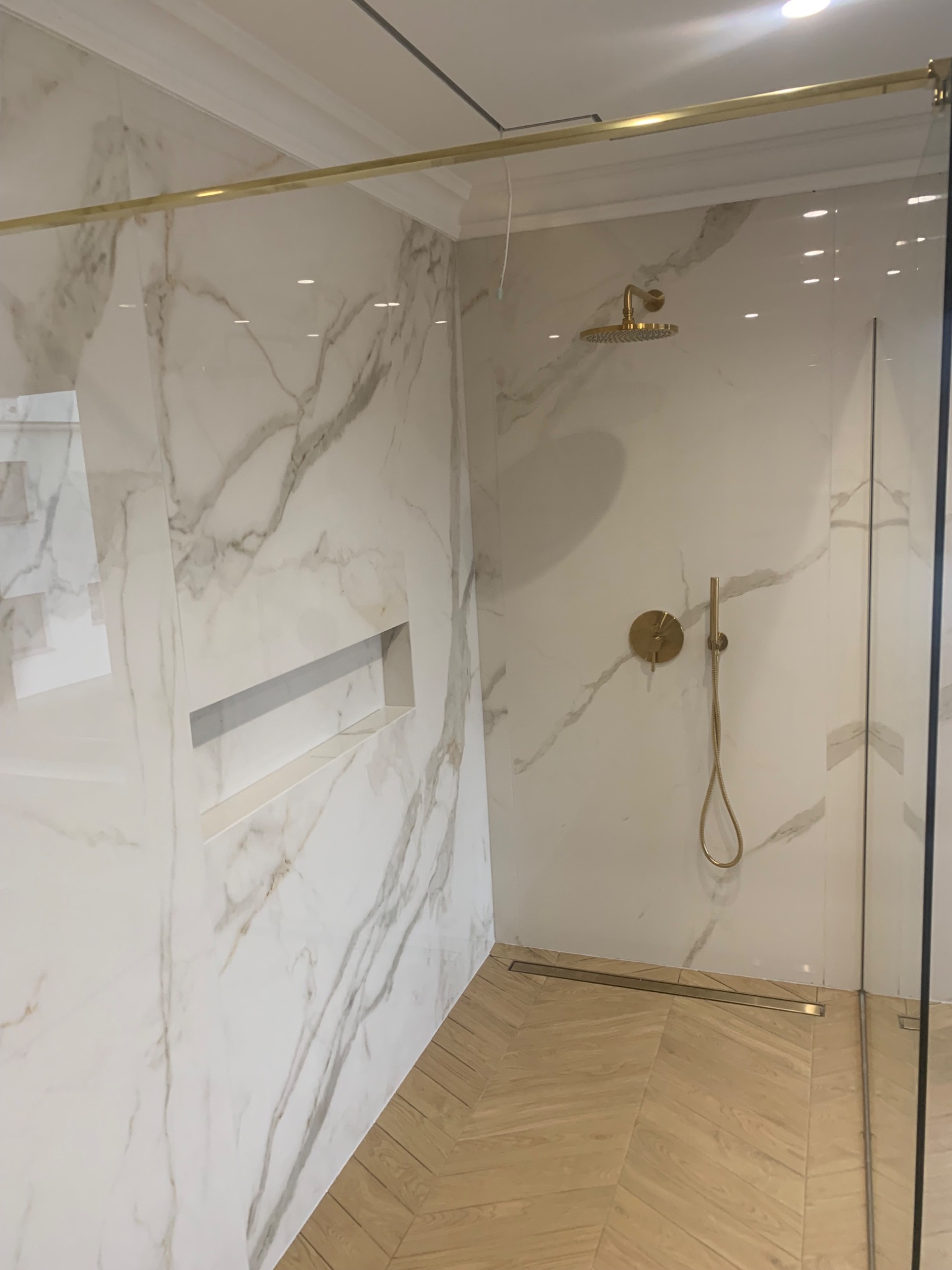 Nowoczesna kabina prysznicowa z marmurowymi płytkami, złotą armaturą i drewnopodobną podłogą ułożoną w jodełkę, widoczna wnęka w ścianie.