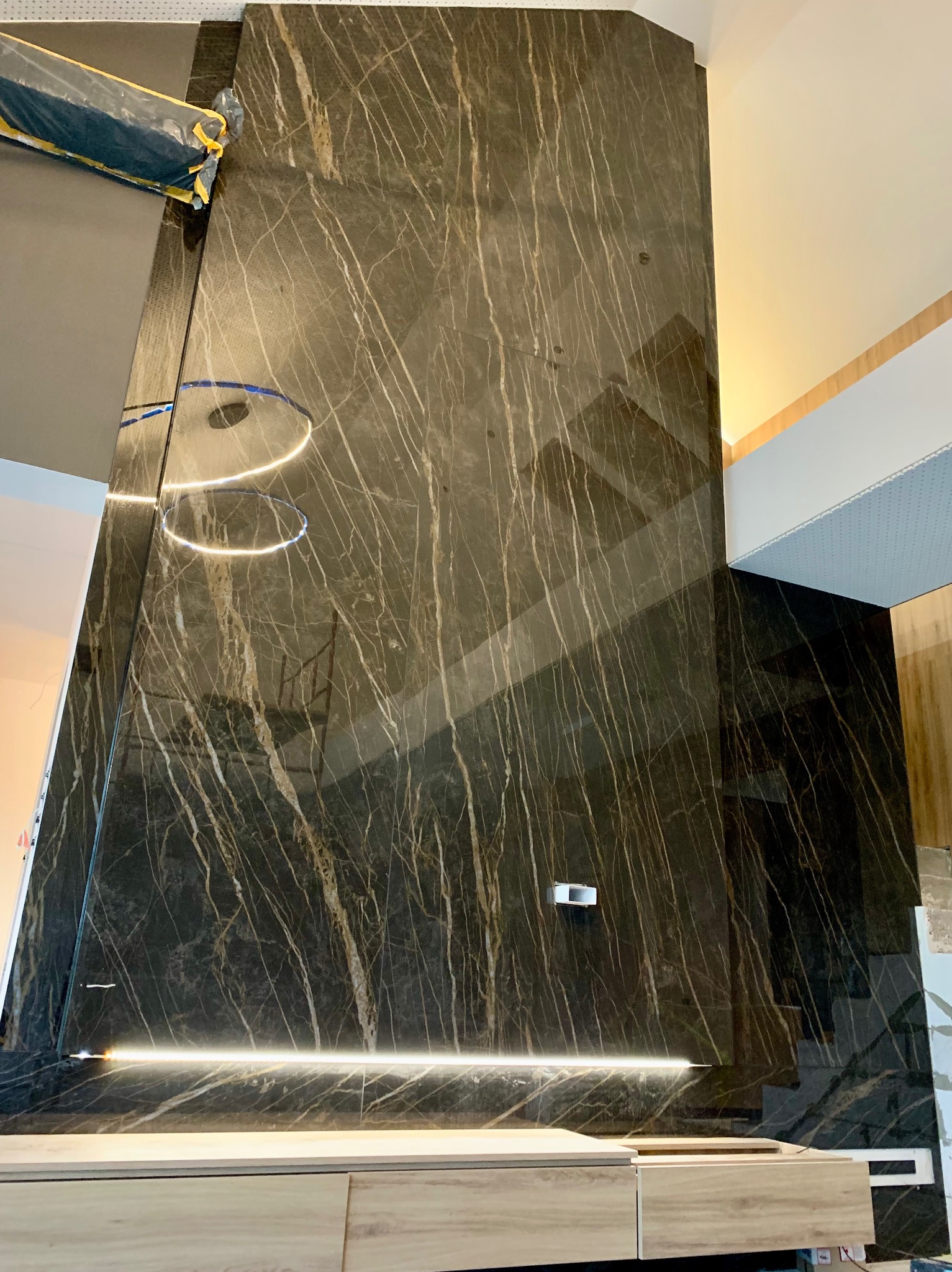 Ściana wyłożona czarnym marmurem z białymi żyłami, oświetlona listwą LED, z widocznymi elementami montażowymi i fragmentami sufitu w trakcie wykończenia.