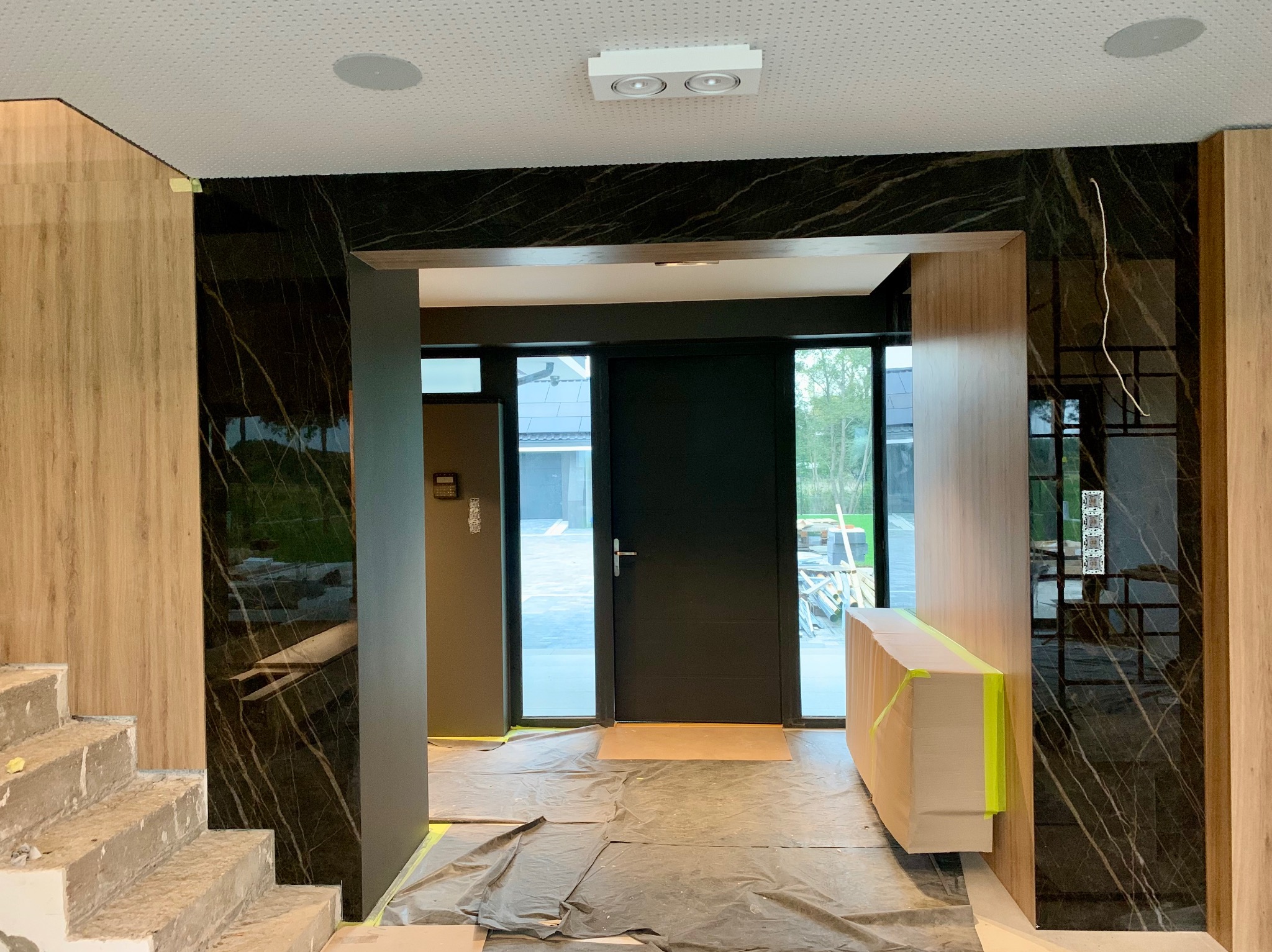 Wnętrze w trakcie prac wykończeniowych: marmurowa ściana, schody w surowym betonie, zabezpieczona folią podłoga, widoczne elementy oświetlenia i drzwi wejściowe.