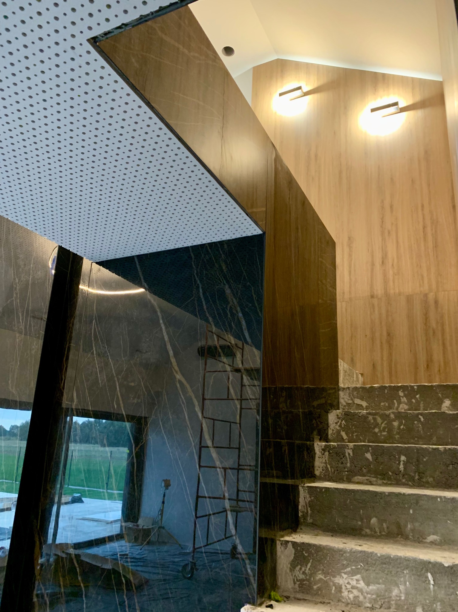 Częściowo wykończona łazienka z marmurowymi płytkami, widoczne schody w stanie surowym, sufit z perforowanej blachy i nowoczesne oświetlenie ścienne.