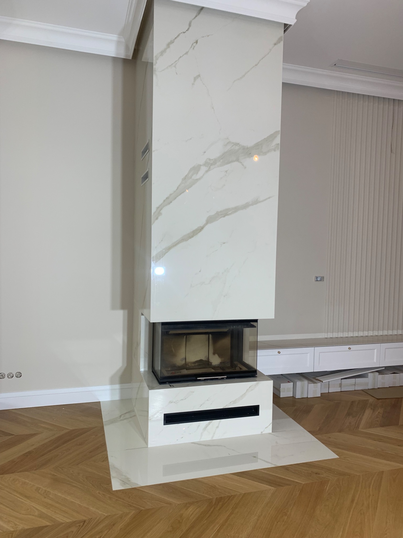 Nowoczesny kominek obudowany białym marmurem z subtelnym, szarym żyłkowaniem, harmonizujący z drewnianą podłogą ułożoną w jodełkę i kremowymi ścianami w tle. Uwagę zwraca minimalistyczny design...