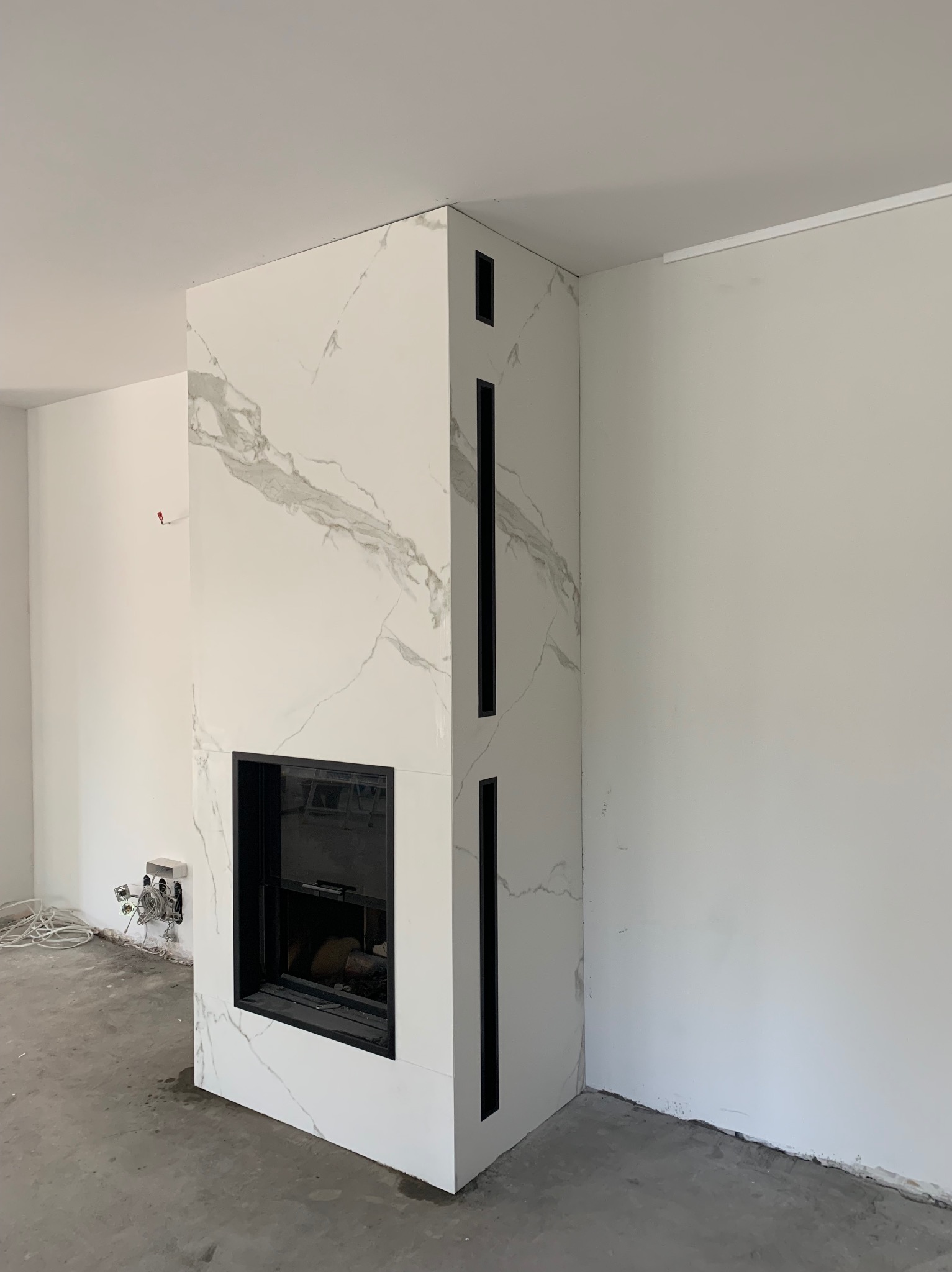 Nowoczesny kominek obudowany białym marmurem z czarnymi akcentami, widoczne niedokończone prace elektryczne i surowy beton na podłodze.