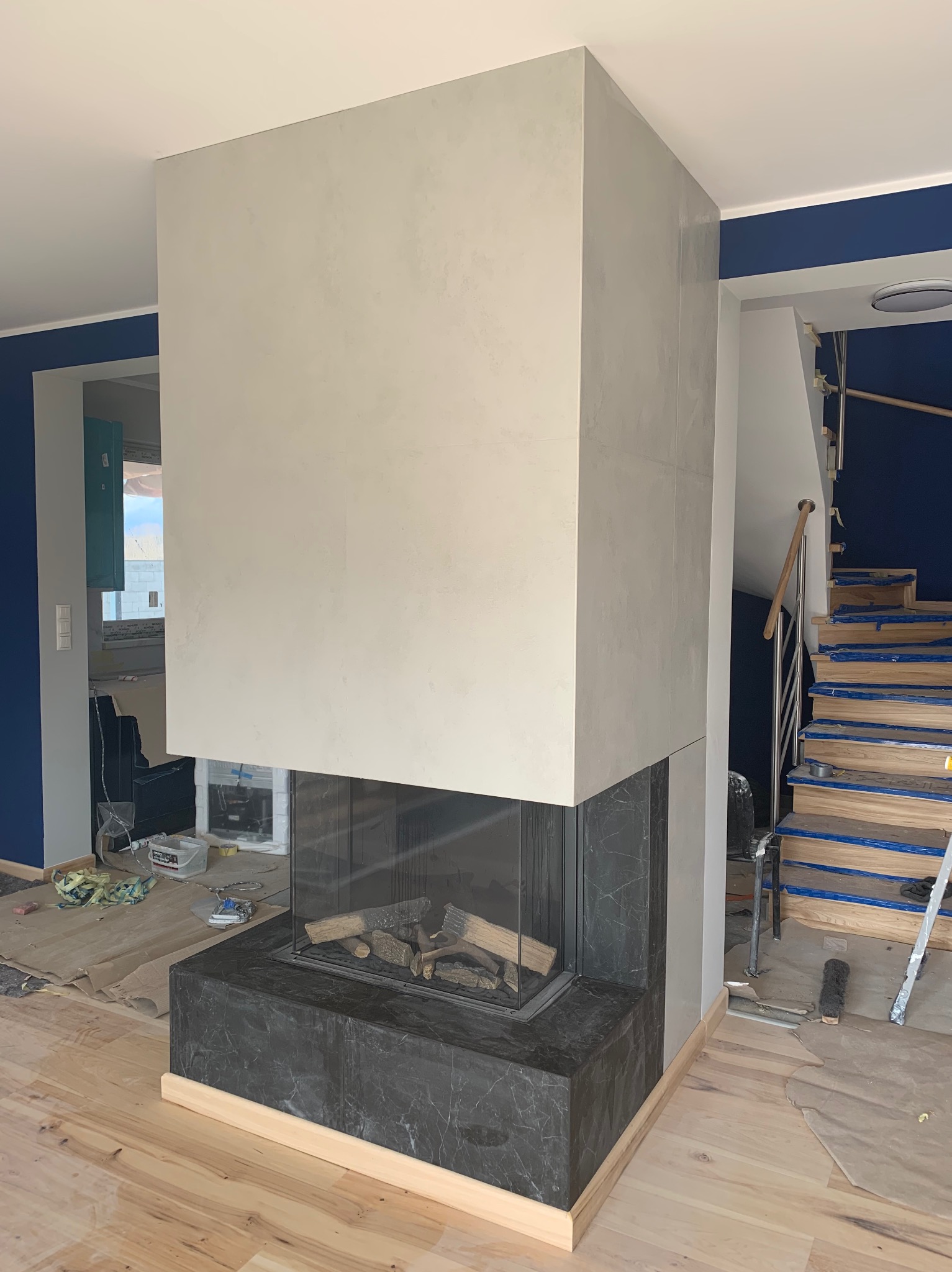 Nowoczesny kominek obudowany jasnoszarą konstrukcją z elementami imitującymi beton, czarny marmurowy cokół, drewniane listwy przypodłogowe, w tle niebieska ściana i schody w trakcie wykończenia.
