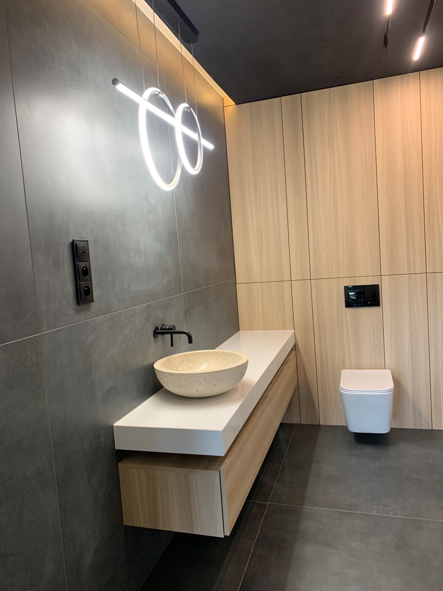 Nowoczesna łazienka z szarymi płytkami, drewnianą zabudową, kamienną umywalką i podwieszaną toaletą, oświetlona podwieszanymi lampami w kształcie okręgów.