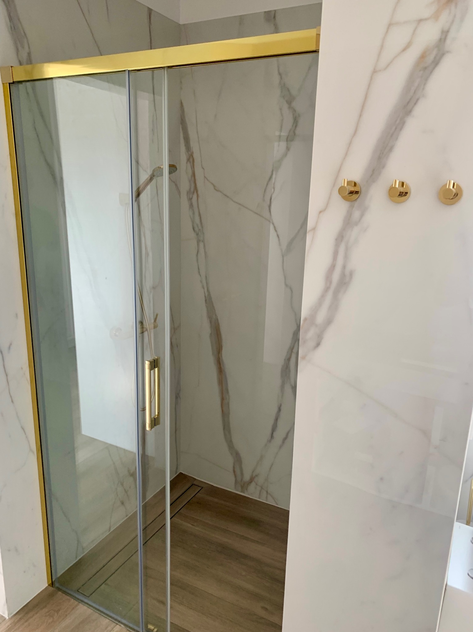 Nowoczesna kabina prysznicowa z przesuwnymi drzwiami, złote wykończenia, marmurowe płytki na ścianach i drewnopodobna podłoga z odpływem liniowym. Widoczne trzy złote wieszaki na ścianie.
