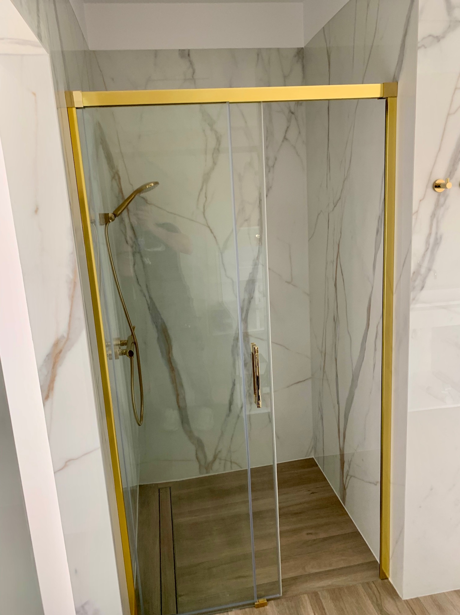 Kabina prysznicowa z przesuwnymi drzwiami, złote profile, marmurowe płytki ścienne z szarymi żyłami, drewnopodobna podłoga z odpływem liniowym.
