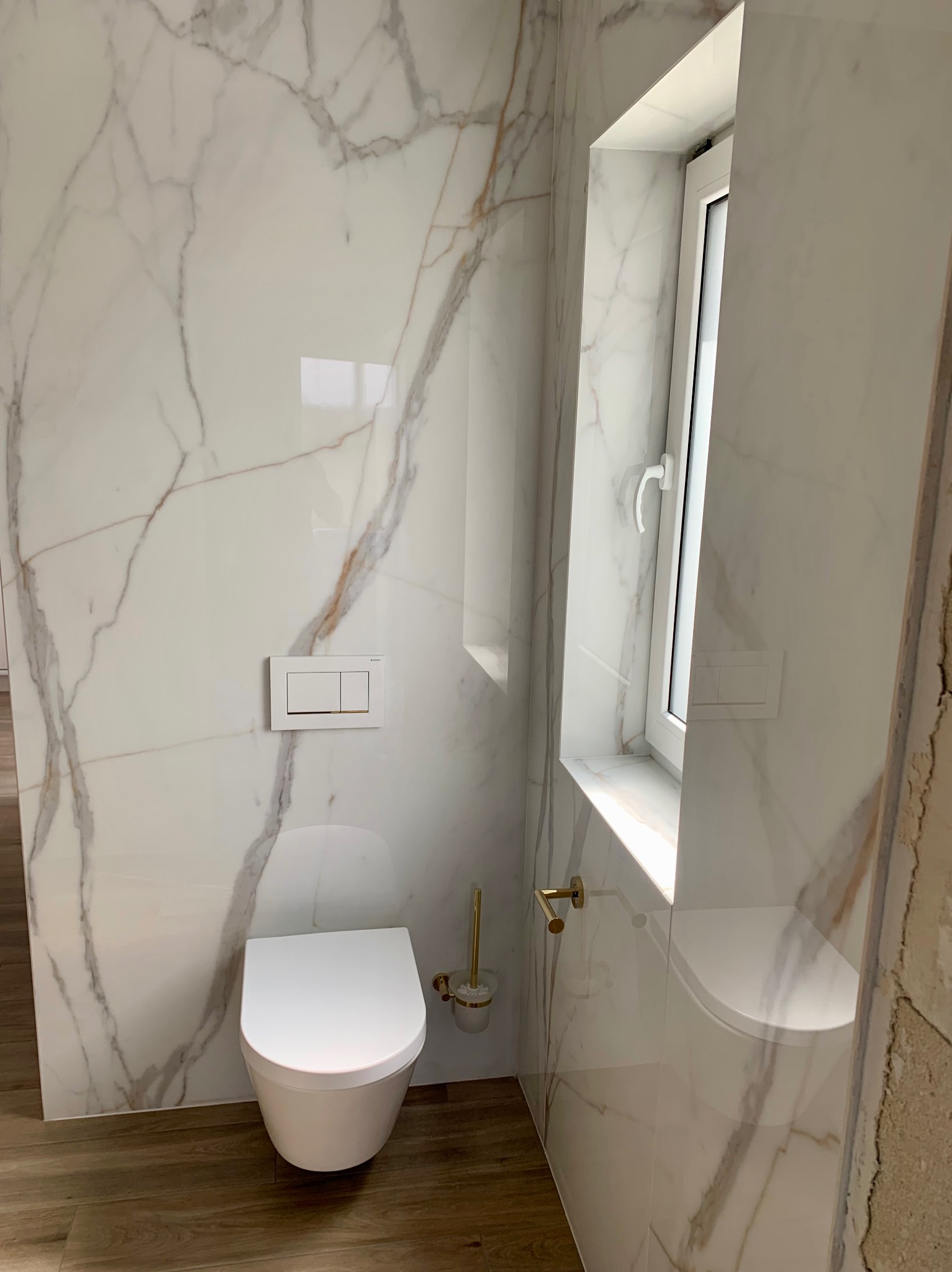 Zawieszona miska WC w nowoczesnej łazience wykończonej płytami imitującymi marmur Calacatta, drewnopodobna podłoga, złote akcenty w postaci uchwytu na szczotkę toaletową i relingu.