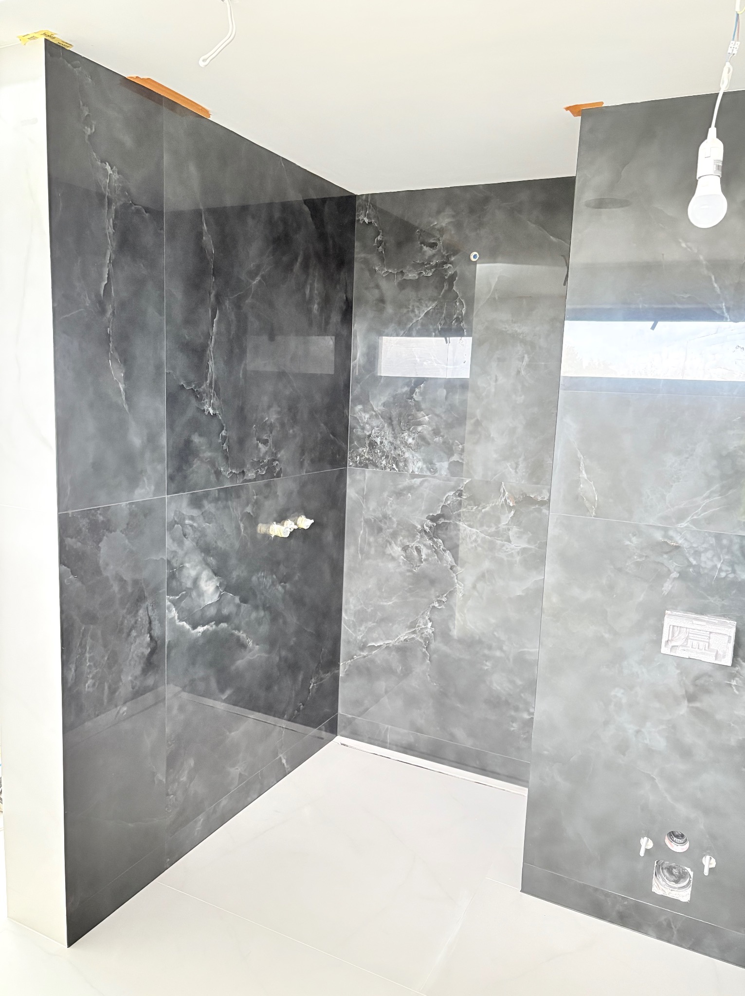 Wykończona łazienka z szarymi płytkami imitującymi marmur na ścianach i białymi płytkami na podłodze. Widoczne przygotowane punkty instalacji wodnej.