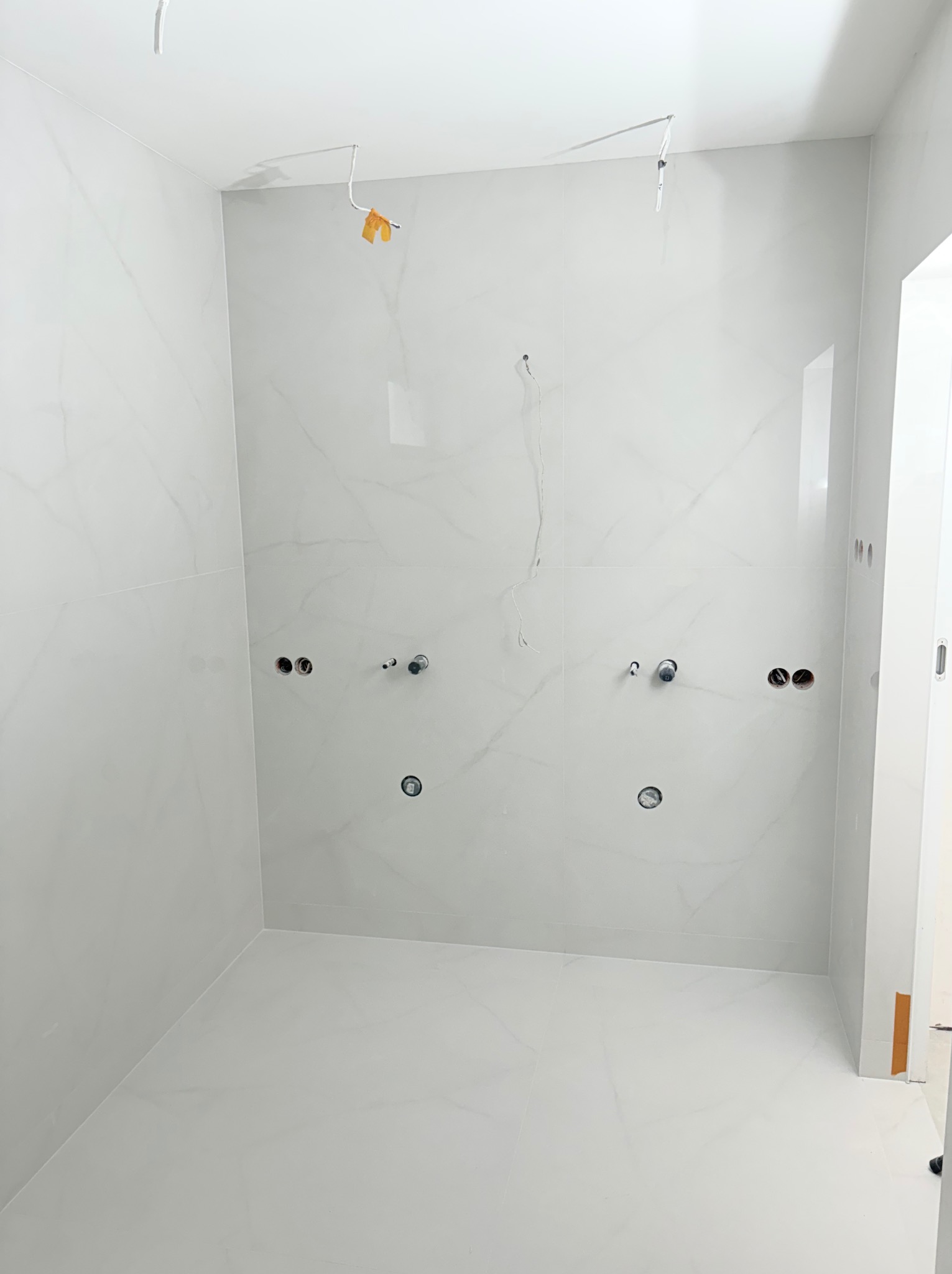 Jasna łazienka z białymi płytkami imitującymi marmur, widoczne przygotowane punkty instalacji wodnej i elektrycznej, surowy stan przed montażem armatury.