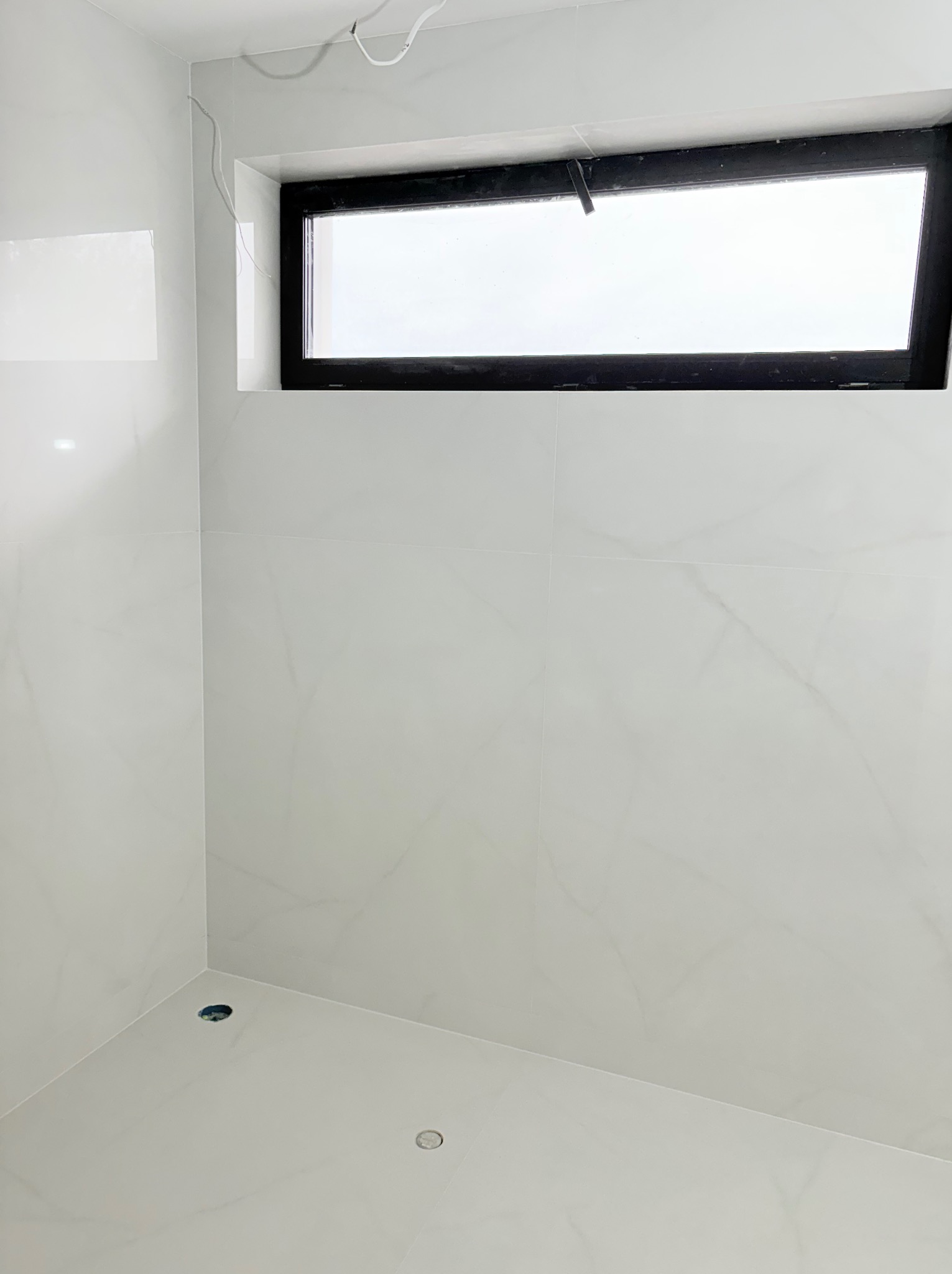 Wykończona łazienka z białymi płytkami imitującymi marmur i czarnym oknem. Widoczne odpływy w podłodze i wystające kable. Minimalistyczny design.