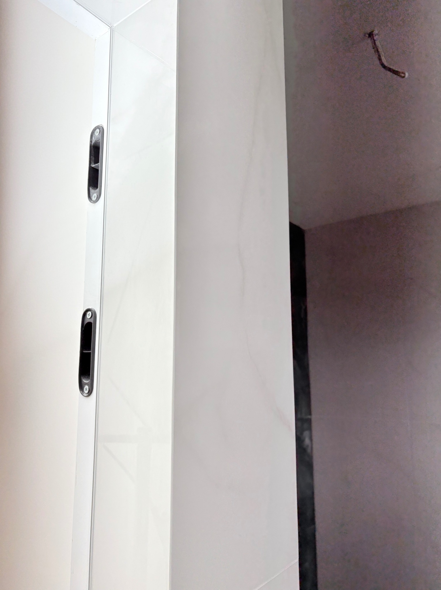 Białe, błyszczące płytki ścienne z zamontowanym profilem i uchwytami w łazience w trakcie wykańczania. Widoczny fragment sufitu z wystającym kablem.