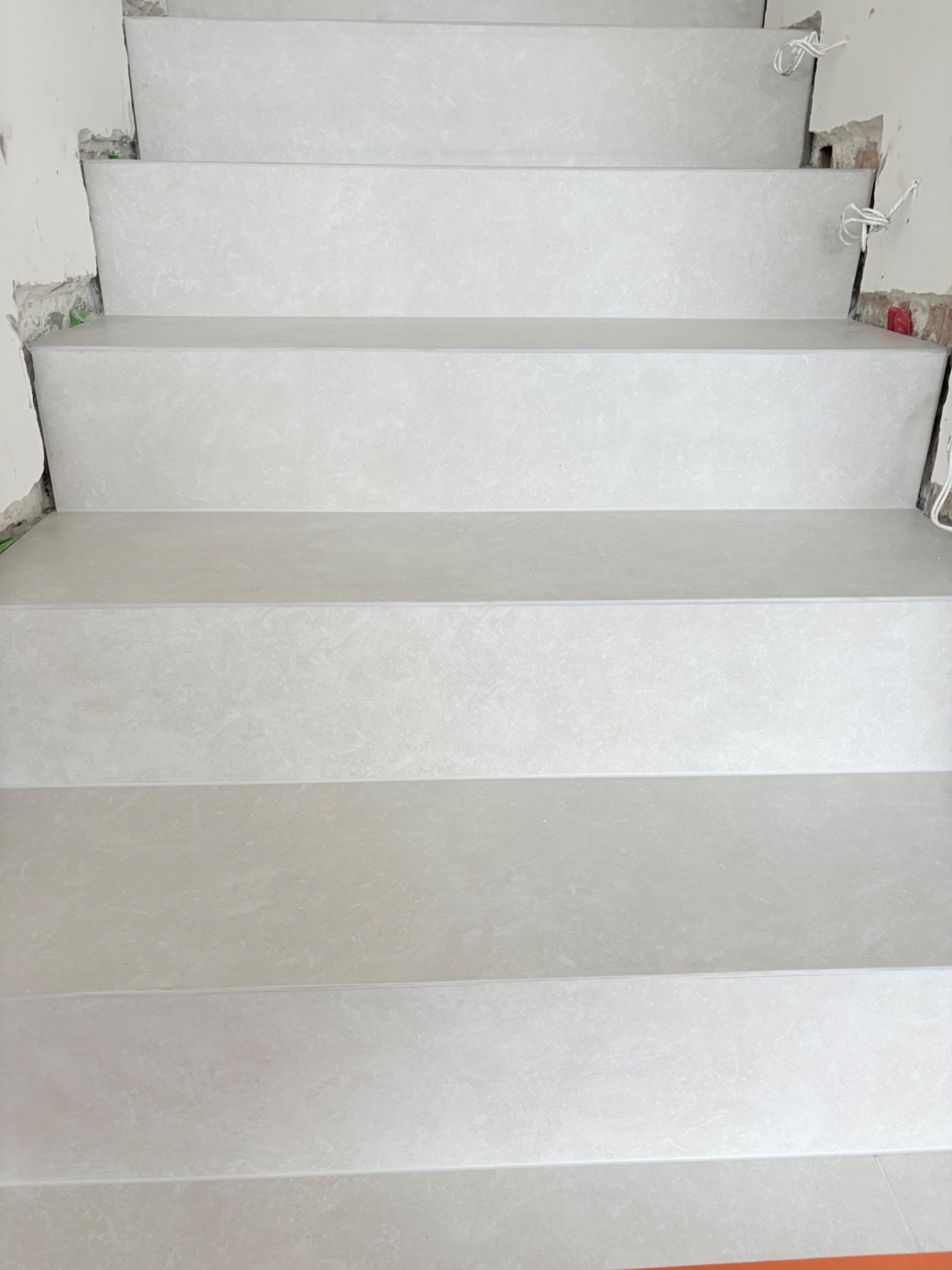 Wykończone schody z jasnych, marmurkowych płytek, widok z góry. Fragment ściany z widocznymi przewodami elektrycznymi. Minimalistyczny design.