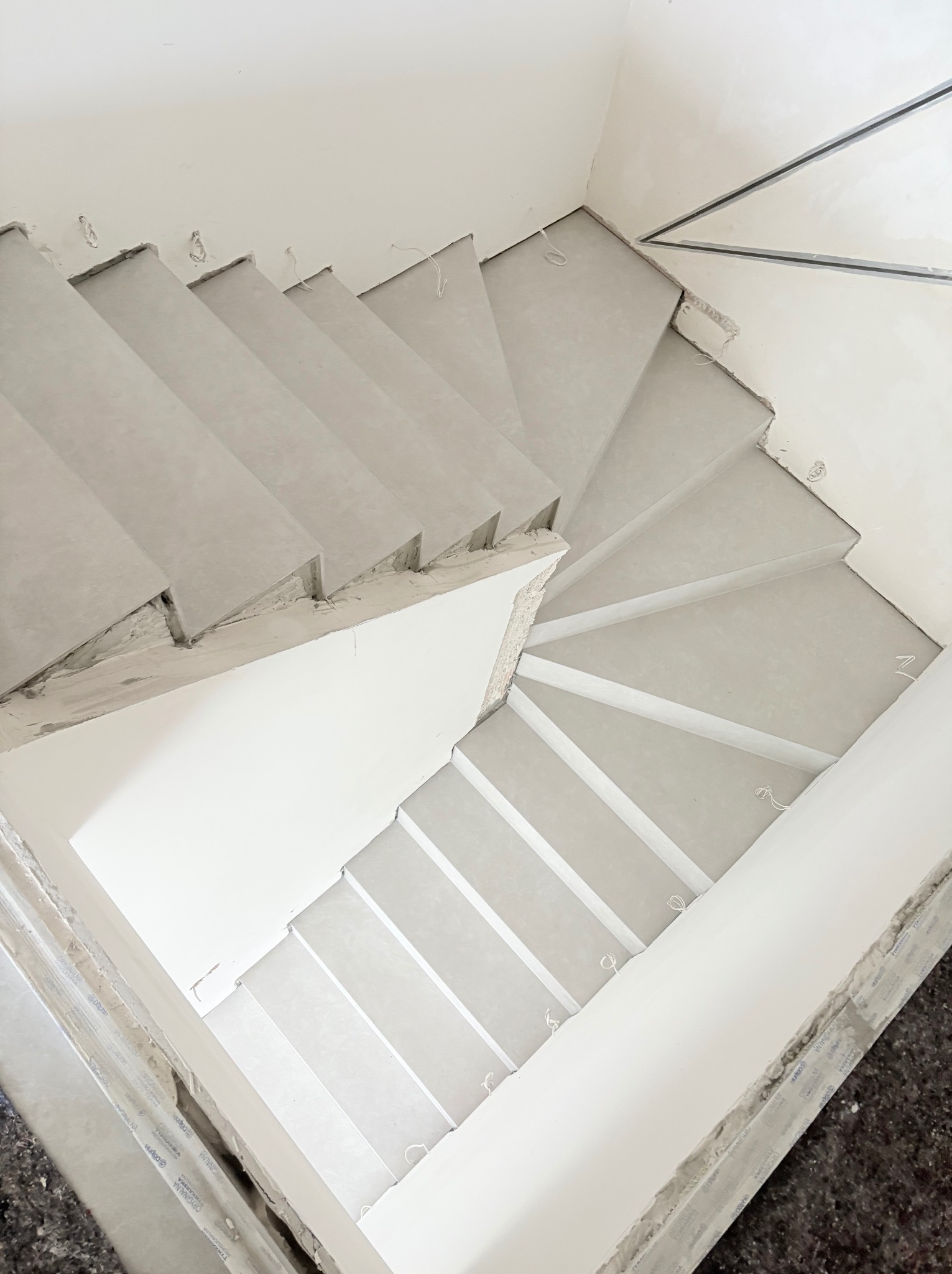Spiralne schody w trakcie wykańczania, widok z góry. Szare stopnie, białe ściany, widoczne kable i niedoskonałości wykończenia. Ujęcie z góry.