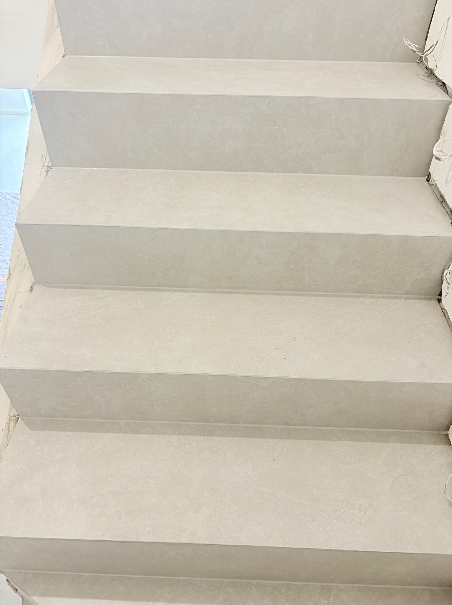 Wykończone schody wewnętrzne z jasnoszarego kamienia, widok z dołu. Minimalistyczny design, surowe wykończenie ścian w tle. Detal kabli elektrycznych.