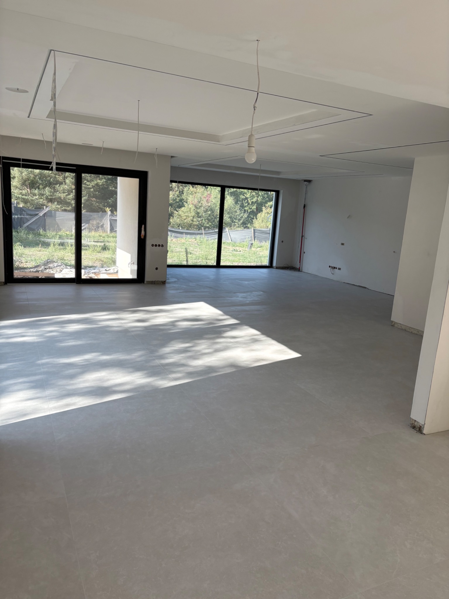 Jasne, przestronne wnętrze z dużymi oknami i szarą podłogą. Widoczne przygotowania do montażu oświetlenia sufitowego. Świeże, minimalistyczne wykończenie.