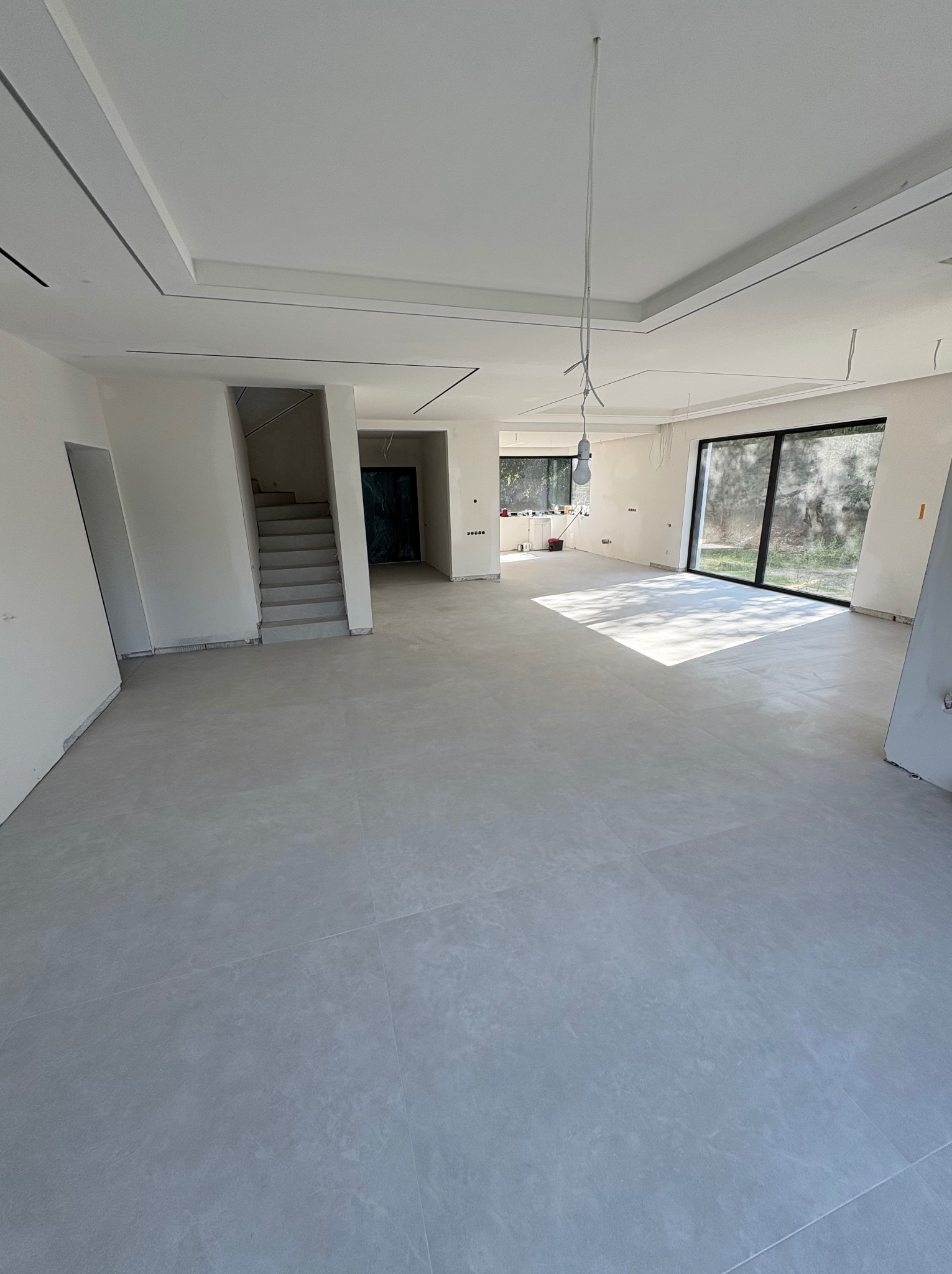 Jasne, przestronne wnętrze w trakcie wykańczania: szare płytki podłogowe, białe ściany, sufit z podwieszanymi elementami. Widoczne okna i schody.