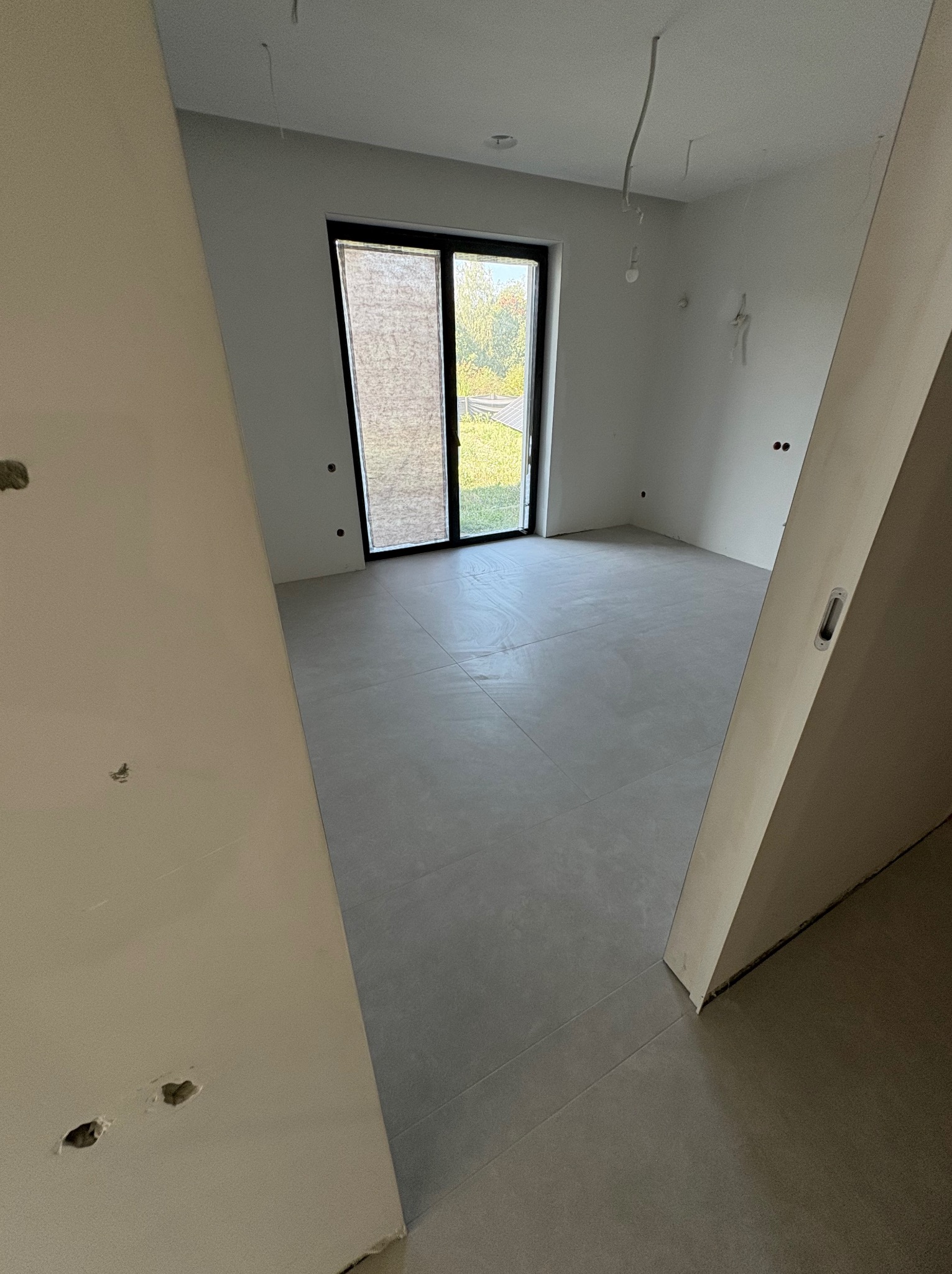 Wnętrze w trakcie prac wykończeniowych: szare płytki podłogowe, białe ściany z widocznymi przewodami elektrycznymi i otworem okiennym z roletą. Widok zza fragmentu ściany.