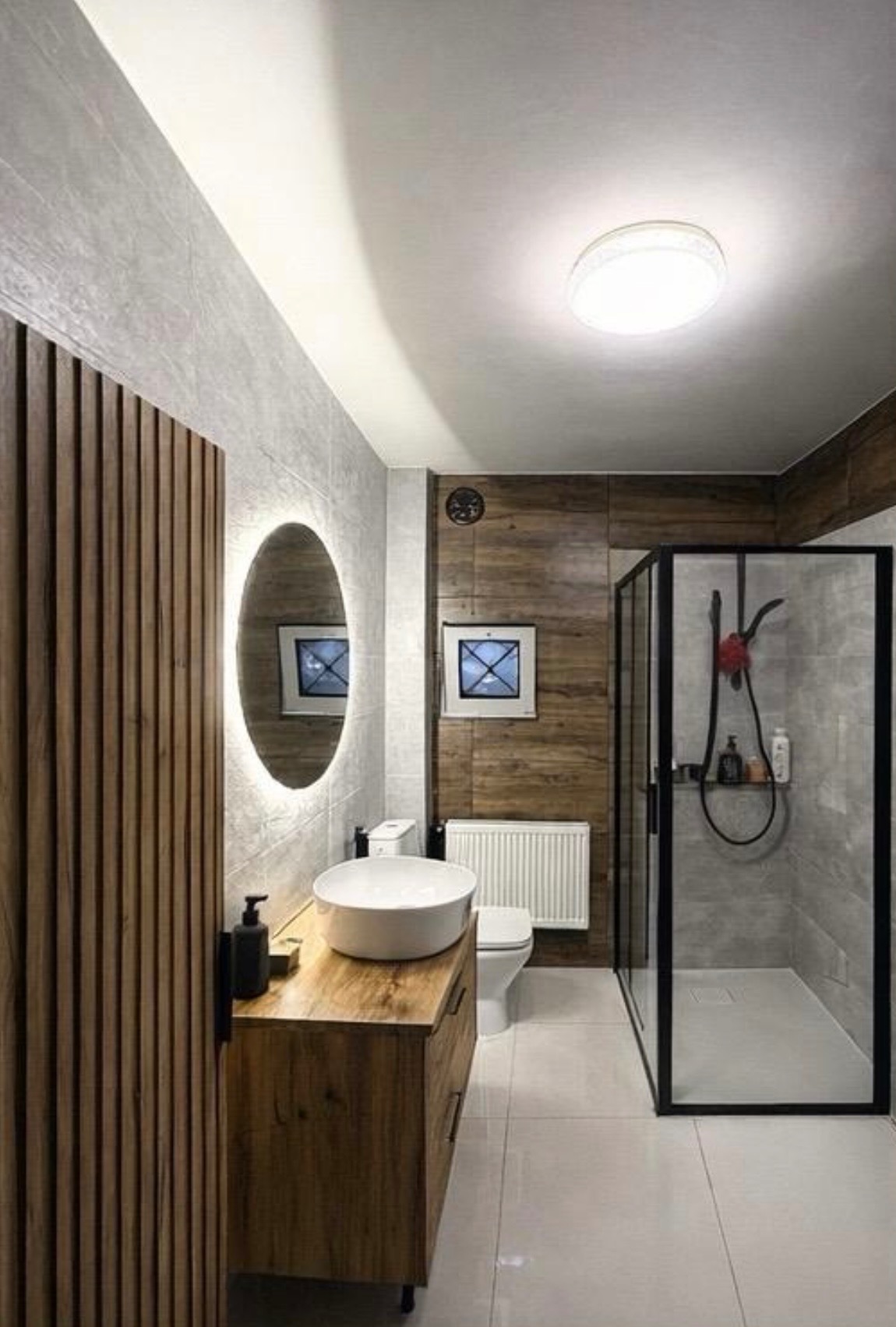 Nowoczesna łazienka z drewnianymi akcentami: szafka pod umywalkę, panel dekoracyjny, kabina prysznicowa z czarną ramą i okrągłe lustro z podświetleniem LED.