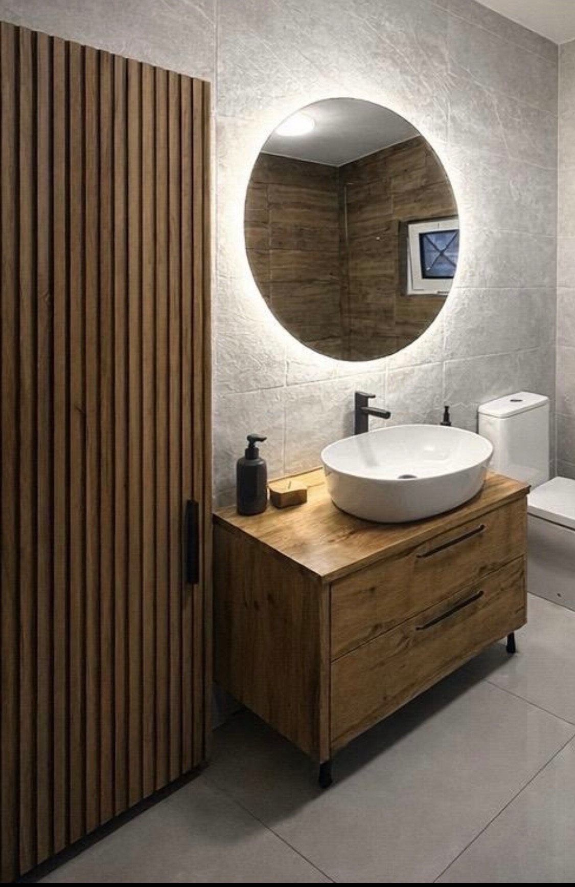 Nowoczesna łazienka z drewnianą szafką pod umywalką, okrągłym lustrem z podświetleniem LED i szarą płytką na ścianach. Minimalistyczny design z akcentami drewna.