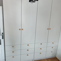 DM Meble - Biała szafa na wymiar z drewnianymi uchwytami, składająca się z części zamykanej drzwiami i części z szufladami, oświetlona wiszącą lampą z trzema kloszami.