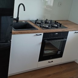 DM Meble - Nowoczesna kuchnia z czarnym zlewem, płytą gazową i piekarnikiem w białych szafkach z drewnianym blatem, obok czarna lodówka. Minimalistyczny design z oknem w tle.