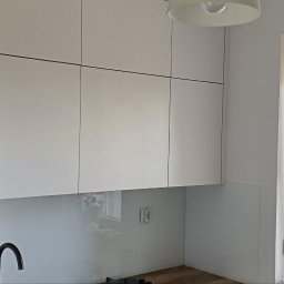 DM Meble - Nowoczesna, jasna kuchnia z białymi szafkami wiszącymi z czarnymi uchwytami i czarną lodówką Samsung. Widok na okno z roletą i miejski krajobraz.