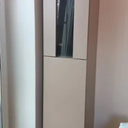 Wysoka szafa z przeszkloną witryną w salonie w Siennicy. Mebel w jasnym odcieniu, minimalistyczny design, widoczne elementy dekoracyjne wewnątrz.