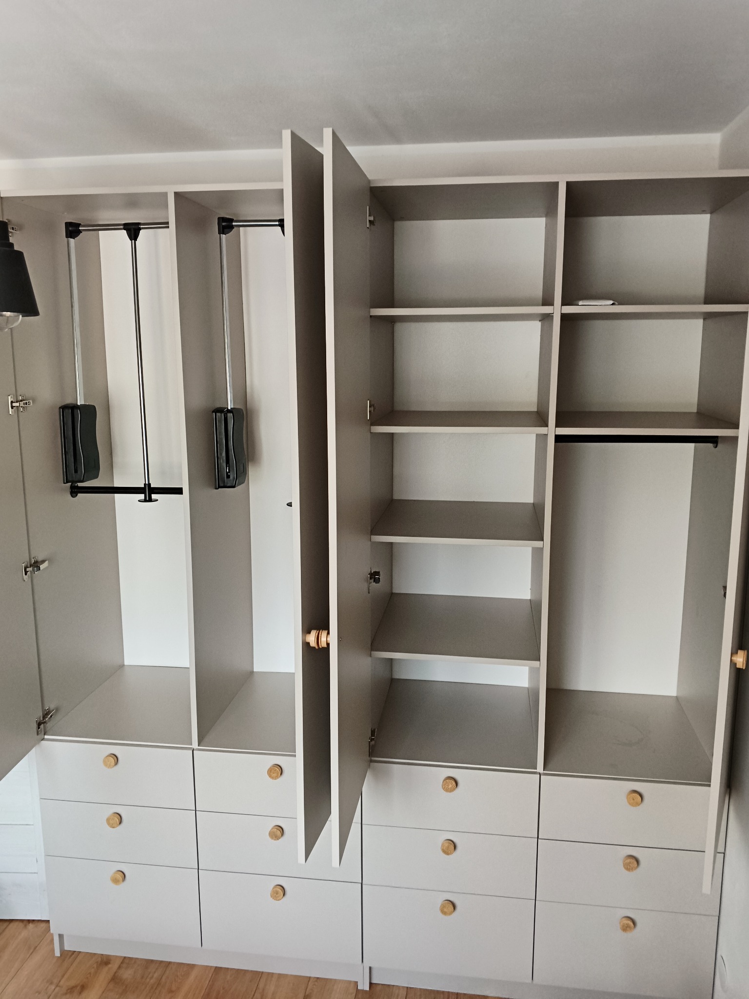 Szara szafa wnękowa z otwartymi drzwiami, widoczne półki, szuflady z okrągłymi drewnianymi uchwytami i wysuwane drążki na ubrania.