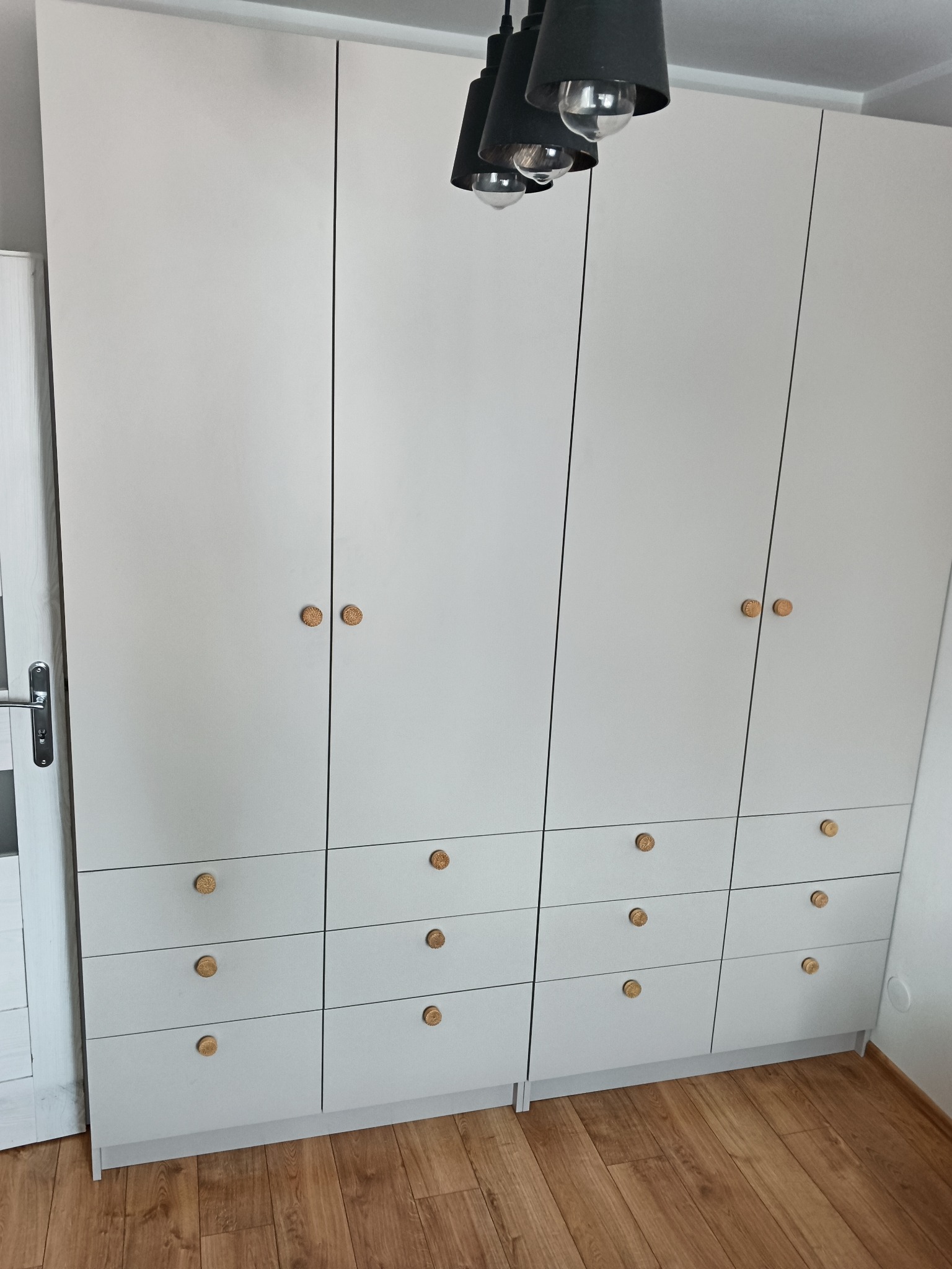 Biała szafa na wymiar z drewnianymi uchwytami, składająca się z części zamykanej drzwiami i części z szufladami, oświetlona wiszącą lampą z trzema kloszami.