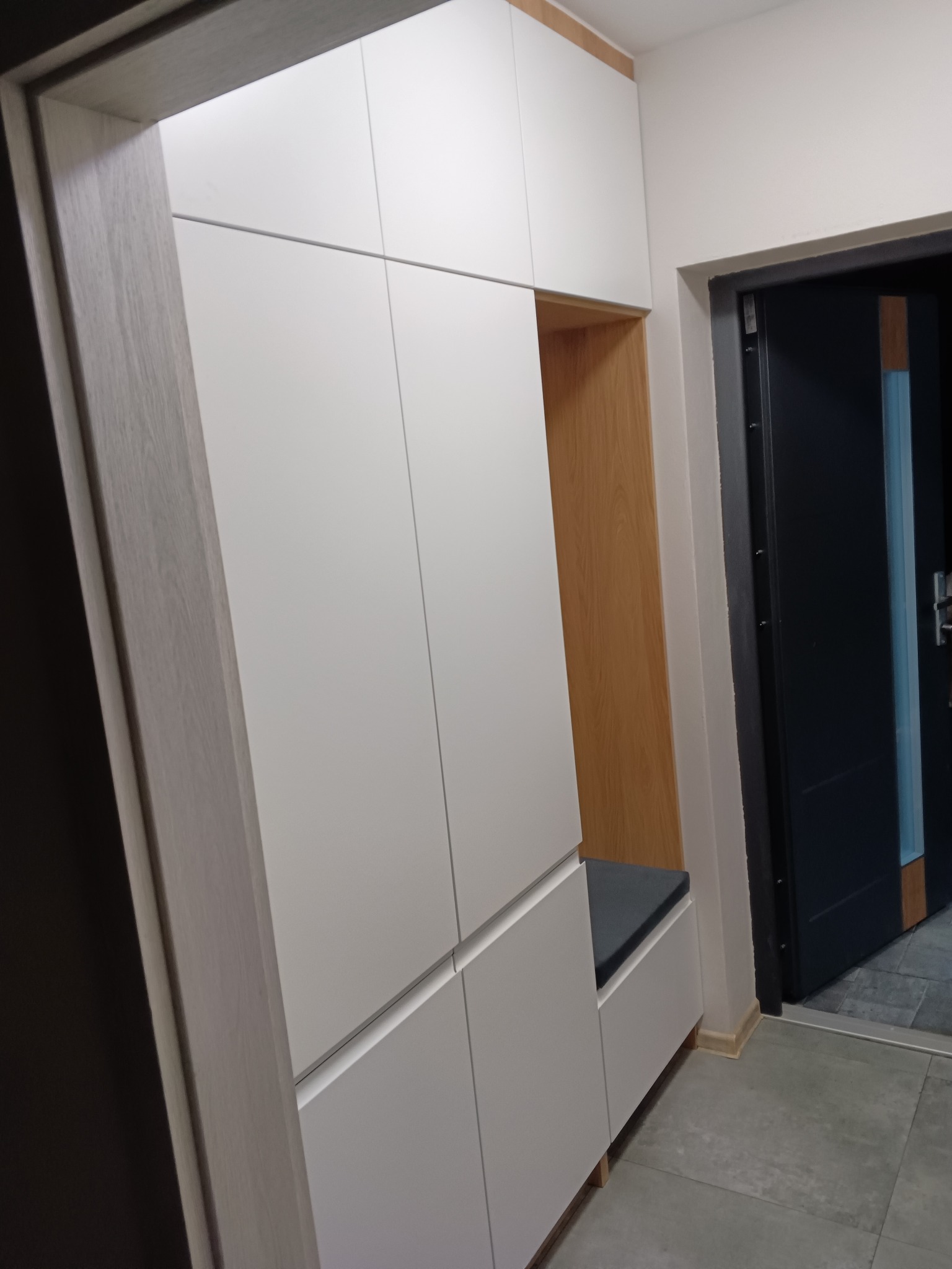 Biała szafa wnękowa z siedziskiem w przedpokoju, widoczna otwarta przestrzeń z drewnianym wykończeniem i czarnym siedziskiem, obok drzwi wejściowe.