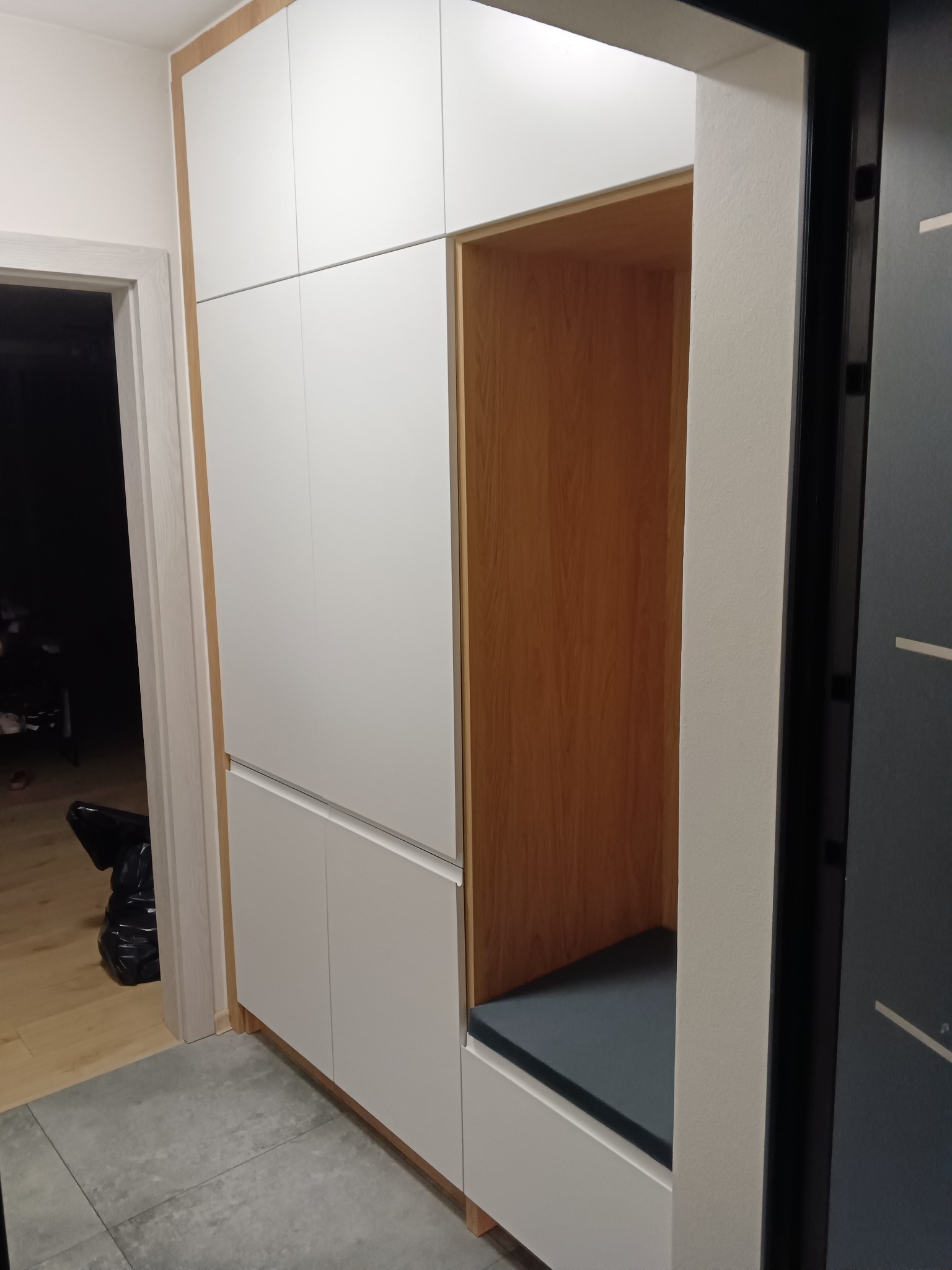 Biała szafa wnękowa z siedziskiem w przedpokoju, wykończona drewnem, szara podłoga.