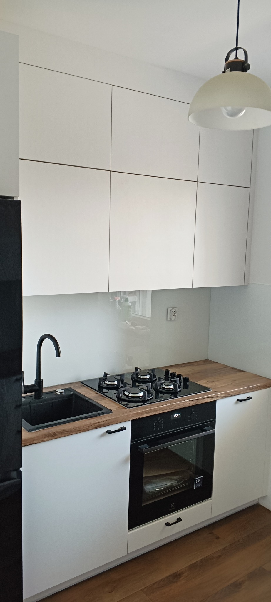 Nowoczesna kuchnia: białe szafki z drewnianym blatem, czarny zlew i płyta gazowa. Minimalistyczny design, lampa wisząca. Widoczne detale wykończenia.