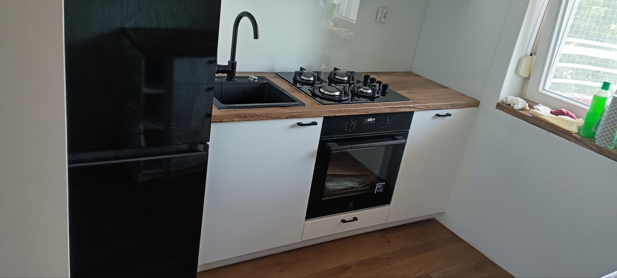 Nowoczesna kuchnia z czarnym zlewem, płytą gazową i piekarnikiem w białych szafkach z drewnianym blatem, obok czarna lodówka. Minimalistyczny design z oknem w tle.