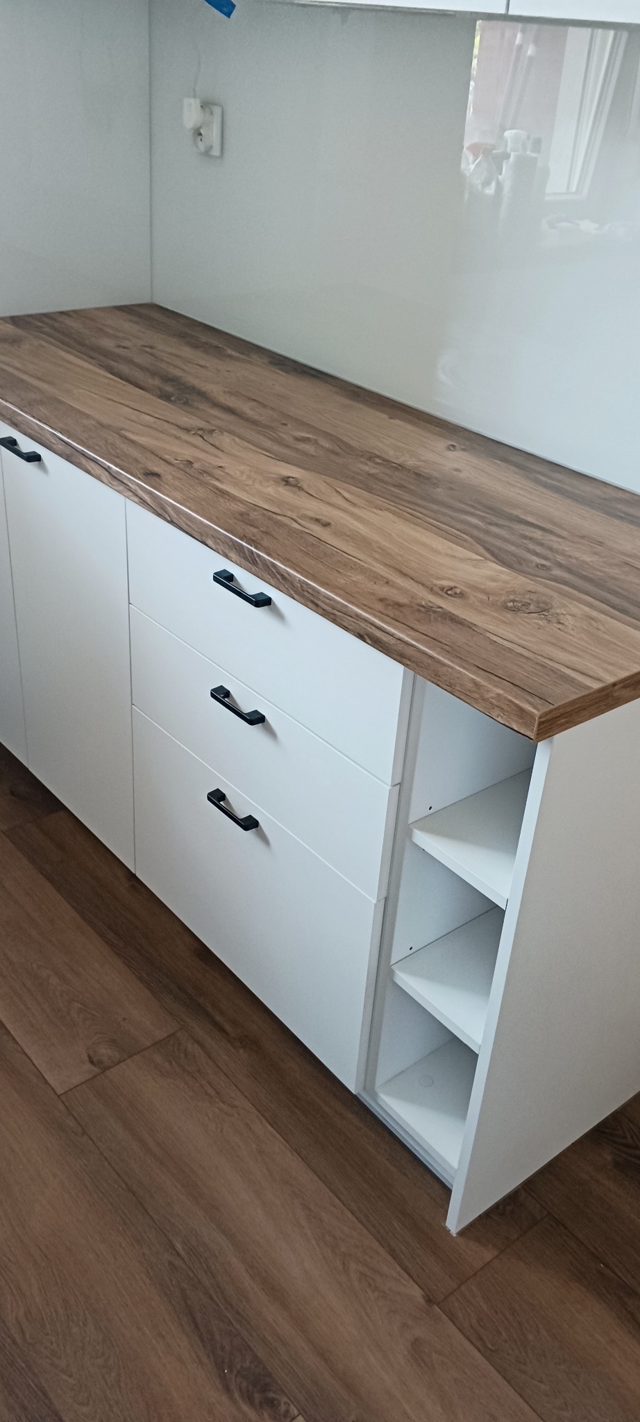 Biała szafka z drewnianym blatem i czarnymi uchwytami, widoczne półki po prawej stronie. Podłoga imitująca drewno. Minimalistyczny design.