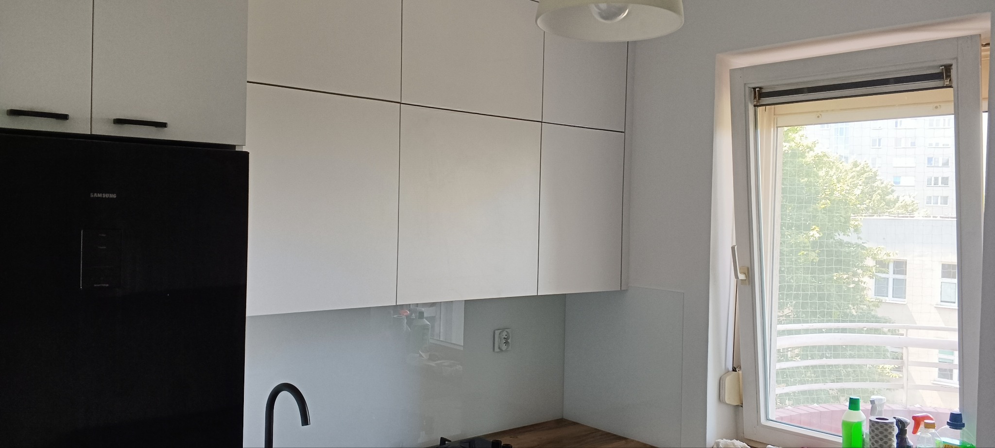 Nowoczesna, jasna kuchnia z białymi szafkami wiszącymi z czarnymi uchwytami i czarną lodówką Samsung. Widok na okno z roletą i miejski krajobraz.