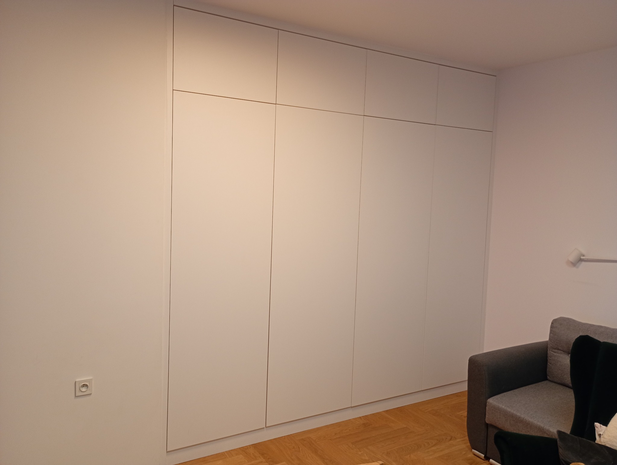 Biała szafa wnękowa z płyty MDF, zlicowana ze ścianą, widoczna w salonie z parkietem i szarą sofą. Minimalistyczny design, brak uchwytów.