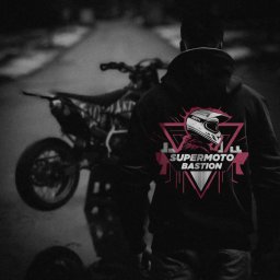 SZOK PRESS POLSKA SPÓŁKA Z OGRANICZONĄ ODPOWIEDZIALNOŚCIĄ - Mężczyzna w bluzie z kapturem z grafiką motocyklową 'Supermoto Bastion' na tle motocykla stojącego na tylnym kole.