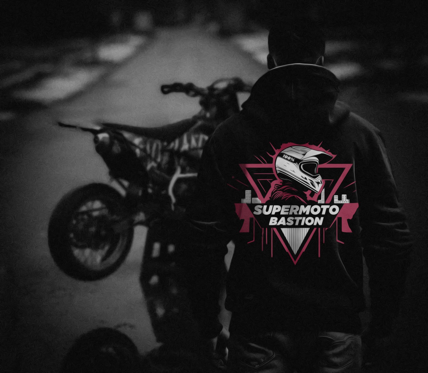 Mężczyzna w bluzie z kapturem z grafiką motocyklową 'Supermoto Bastion' na tle motocykla stojącego na tylnym kole.