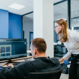 Architektura wnętrz biura: mężczyzna pracuje przy komputerze z widocznym projektem budowlanym, kobieta w okularach pochyla się, obserwując ekran.