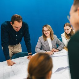 Małro Projekt sp. z o.o. - Zespół architektów analizuje plany budynku na dużym stole w biurze z niebieską ścianą, kobieta w okularach uśmiecha się wskazując na szczegóły na planie.