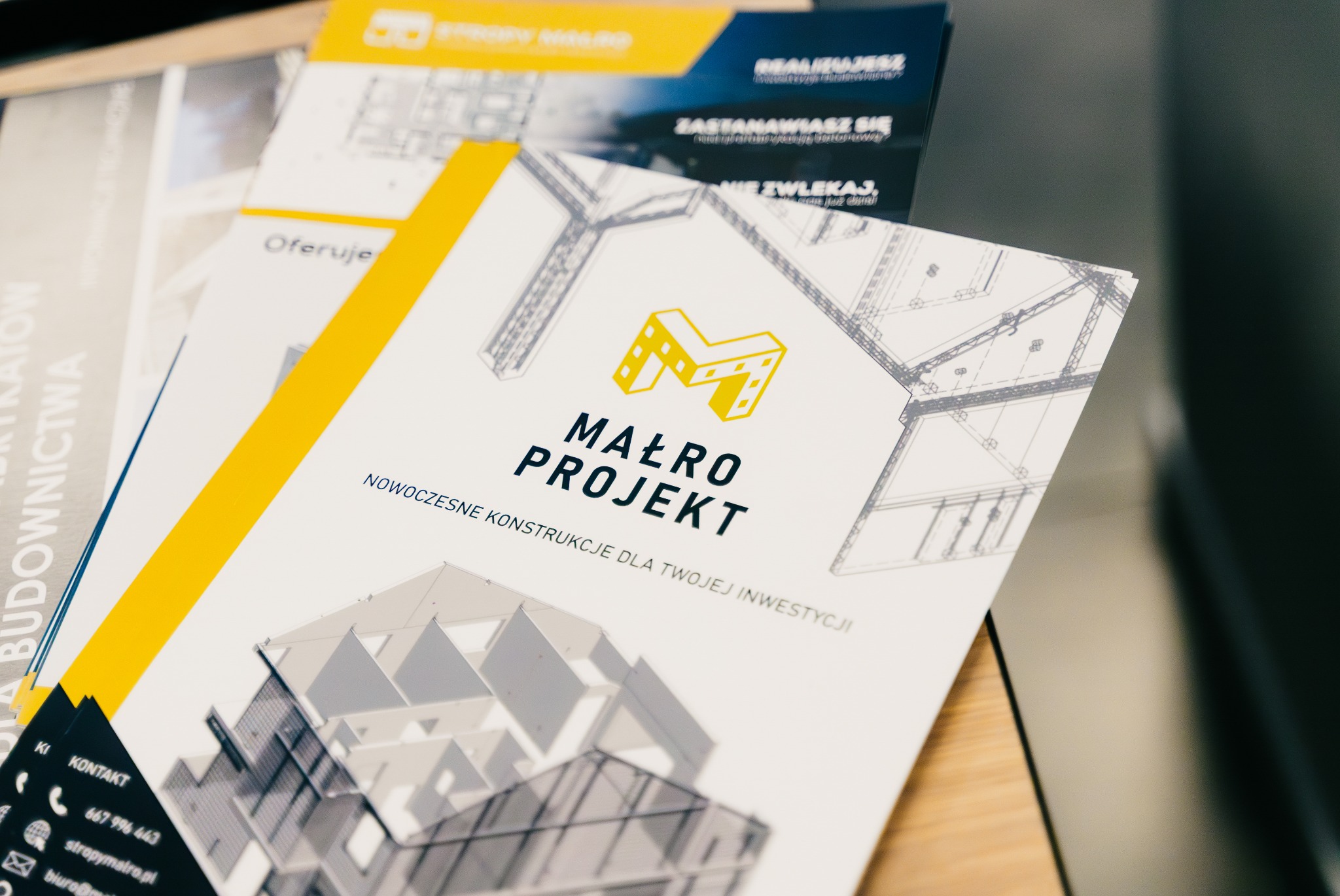 Stos ulotek reklamowych firmy budowlanej z żółtym paskiem i logo, prezentujących wizualizację nowoczesnego budynku z elementami konstrukcji stalowych.