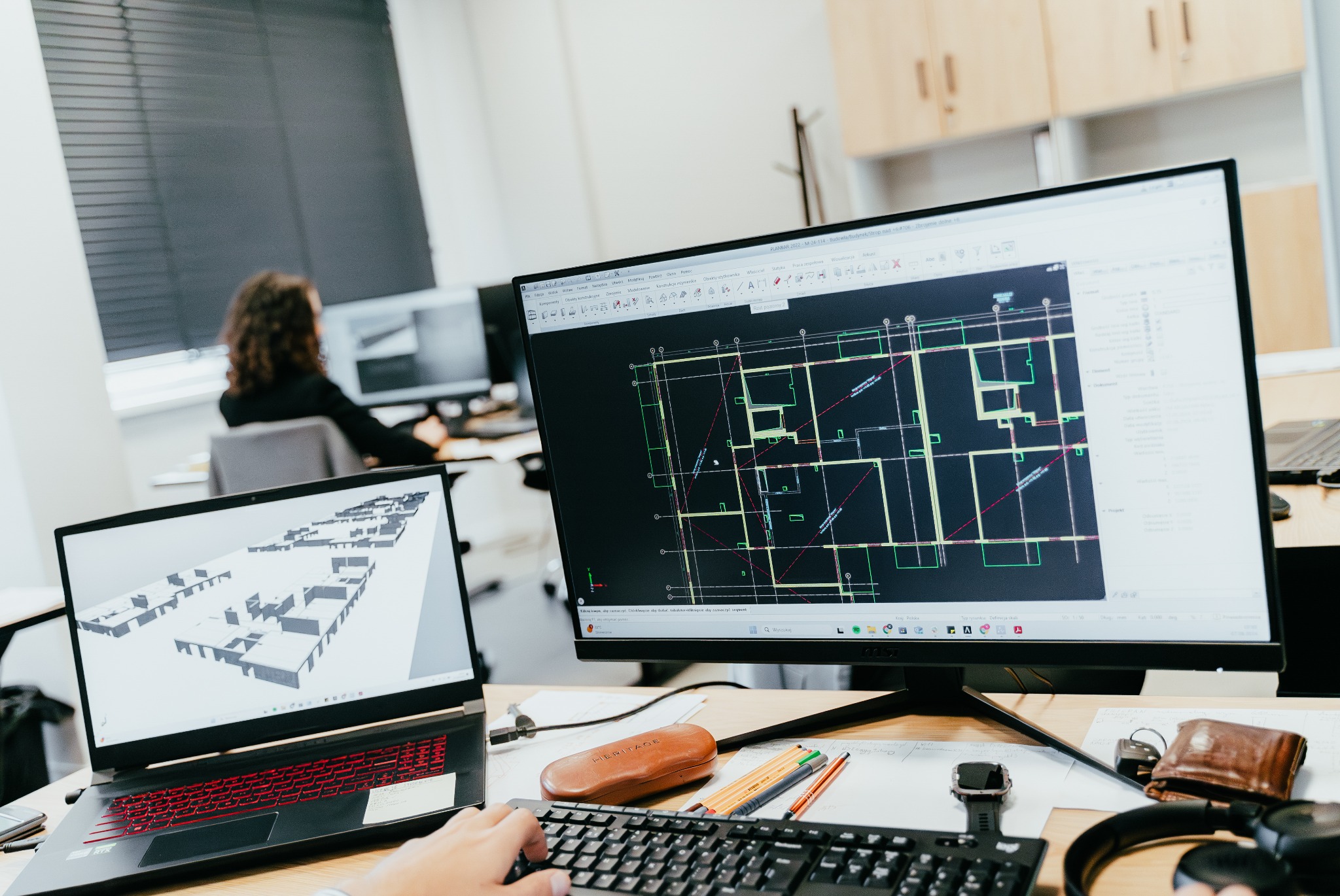 Biuro architektoniczne: widok na biurko z dwoma ekranami, na jednym z nich widoczny plan budynku, a na drugim model 3D, obok leżą okulary, długopisy, notatki i słuchawki.
