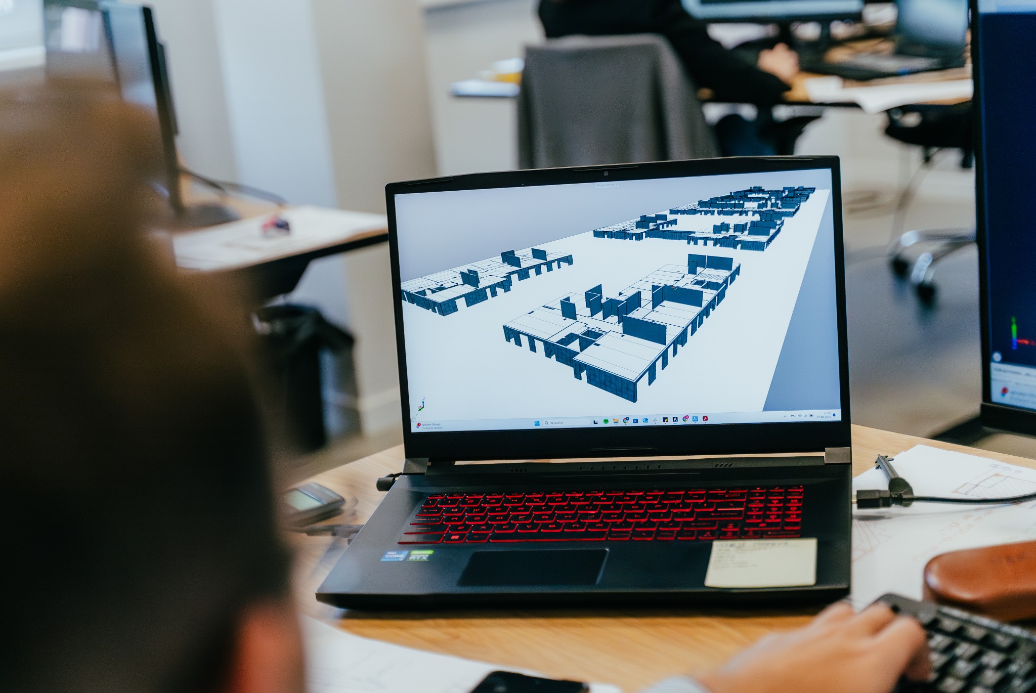 Laptop z czerwonym podświetleniem klawiatury wyświetla trójwymiarowy model architektoniczny budynku z płaskim dachem. Widoczny fragment dłoni osoby pracującej na klawiaturze. W tle biurko z innymi...